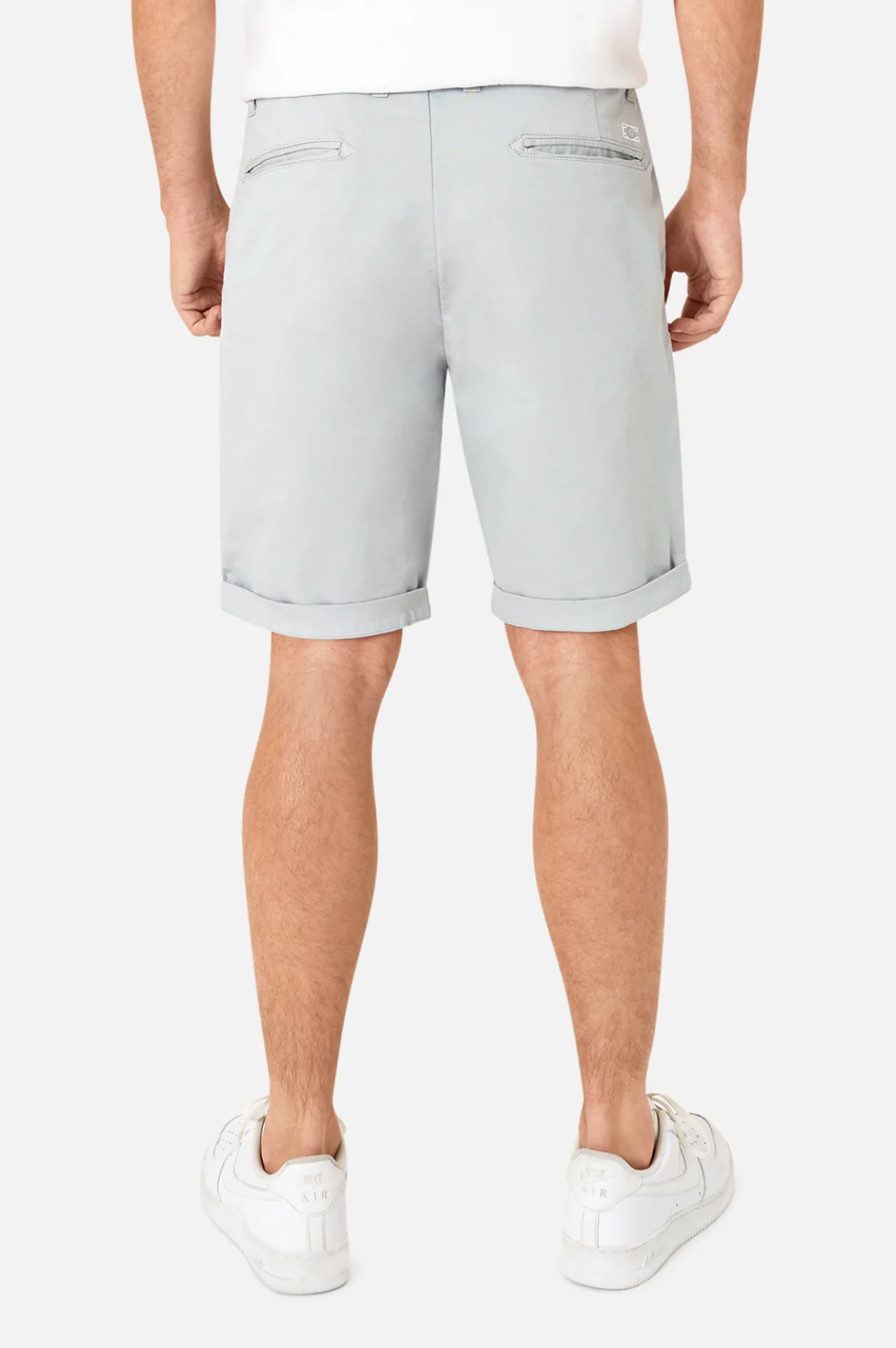 The person is wearing Industrie The Rinse Drifter Short, light gray cotton with relaxed fit and rolled-up hems, paired with white sneakers and a simple white shirt. They stand with their back to the camera, showcasing the shorts' two back pockets.