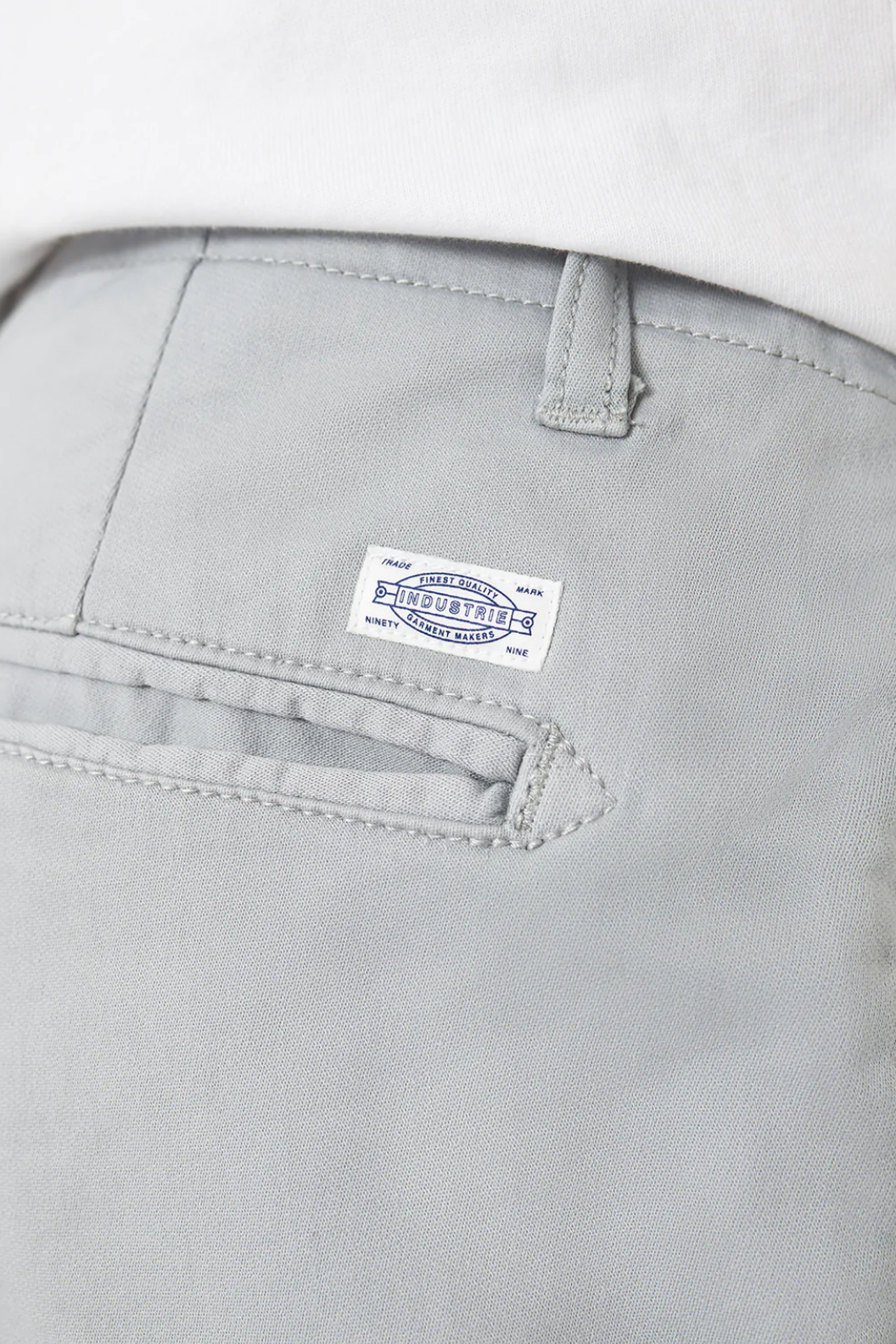 Close-up of light gray Industrie The Rinse Drifter Shorts with a small white label and blue text above the back pocket, worn by someone whose white shirt is partly visible at the top.