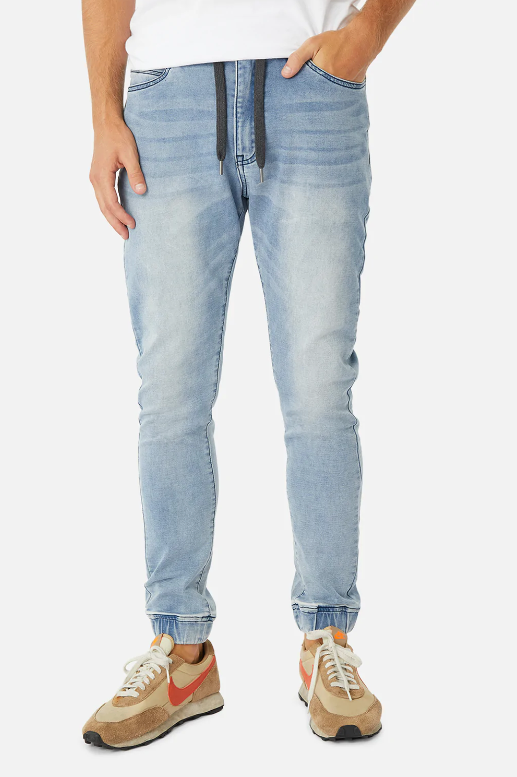 Wearing Industrie The Drifter Denim Pant, a person stands with hands in pockets. The light blue, relaxed-fit jeans feature a drawstring waist and cuffed ankles. They also wear a white shirt and beige sneakers with orange accents.