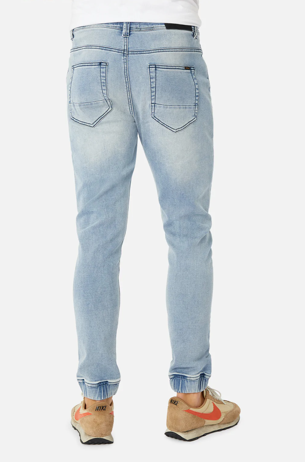 A person wears Industrie The Drifter Denim Pant, featuring a relaxing light blue jogger style with back pockets and elastic cuffs, paired with tan and orange sneakers. They stand with their back turned, exuding a casual vibe against a plain white background.