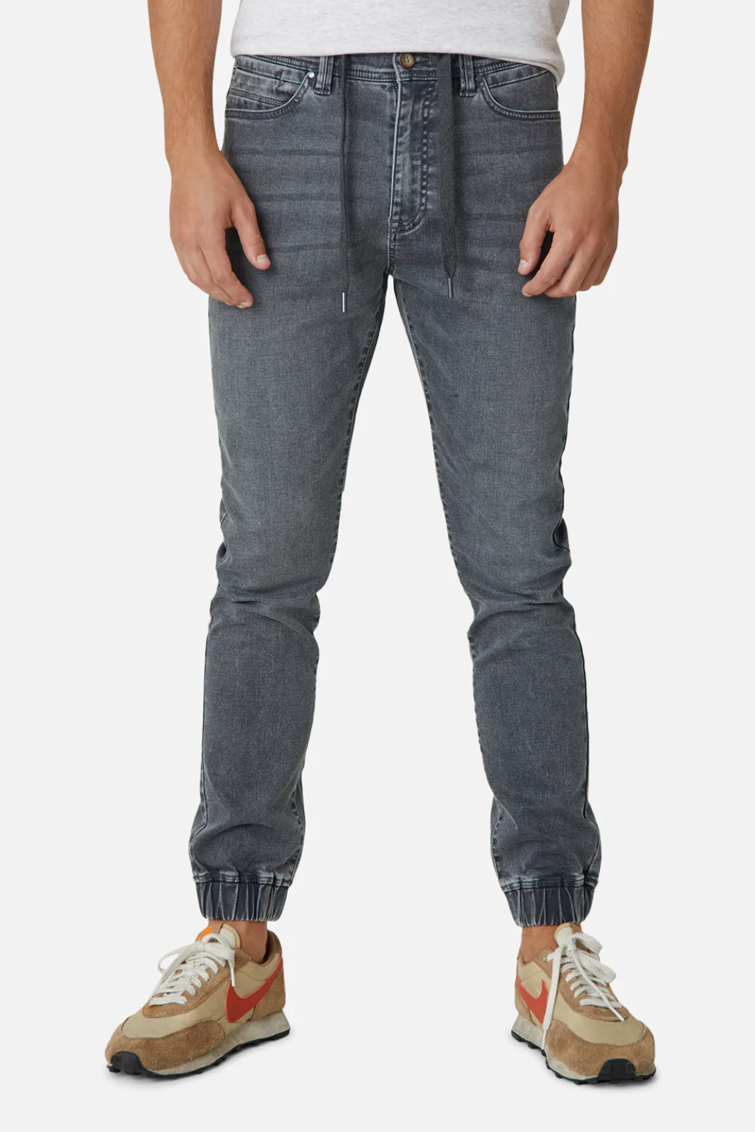 A person is standing in a pair of Industrie The Drifter Denim Pants with a relaxed fit and elastic ankles, featuring a white drawstring. Paired with beige sneakers adorned with red accents and white laces, they wear a white shirt, though only the lower portion is visible.