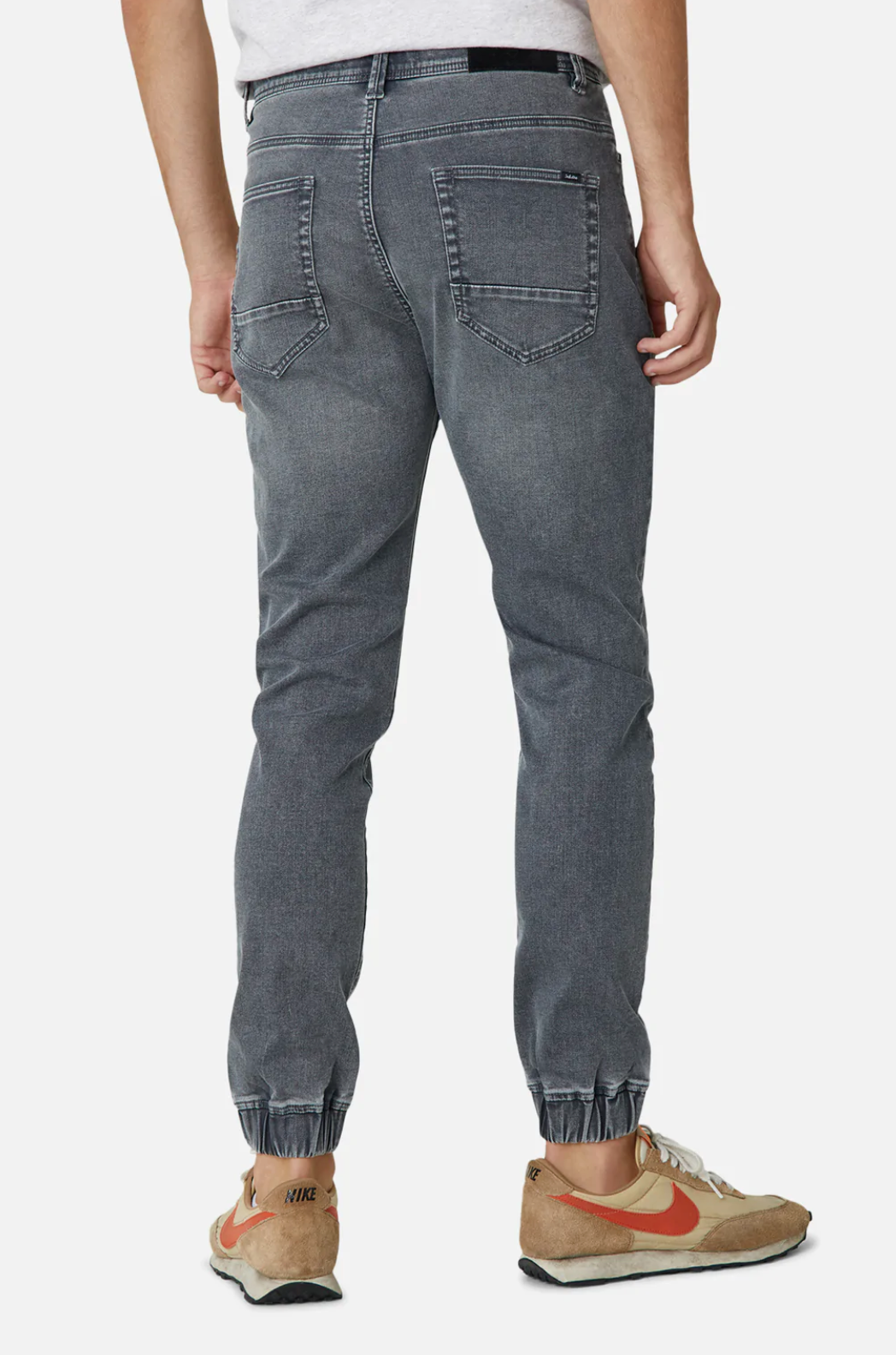 Rear view of a person wearing Industrie The Drifter Denim Pant with a relaxed fit, paired with beige and orange Nike sneakers. The jeans feature back pockets and cuffed legs for added style. The person is also wearing a white shirt tucked into the comfortable jeans.