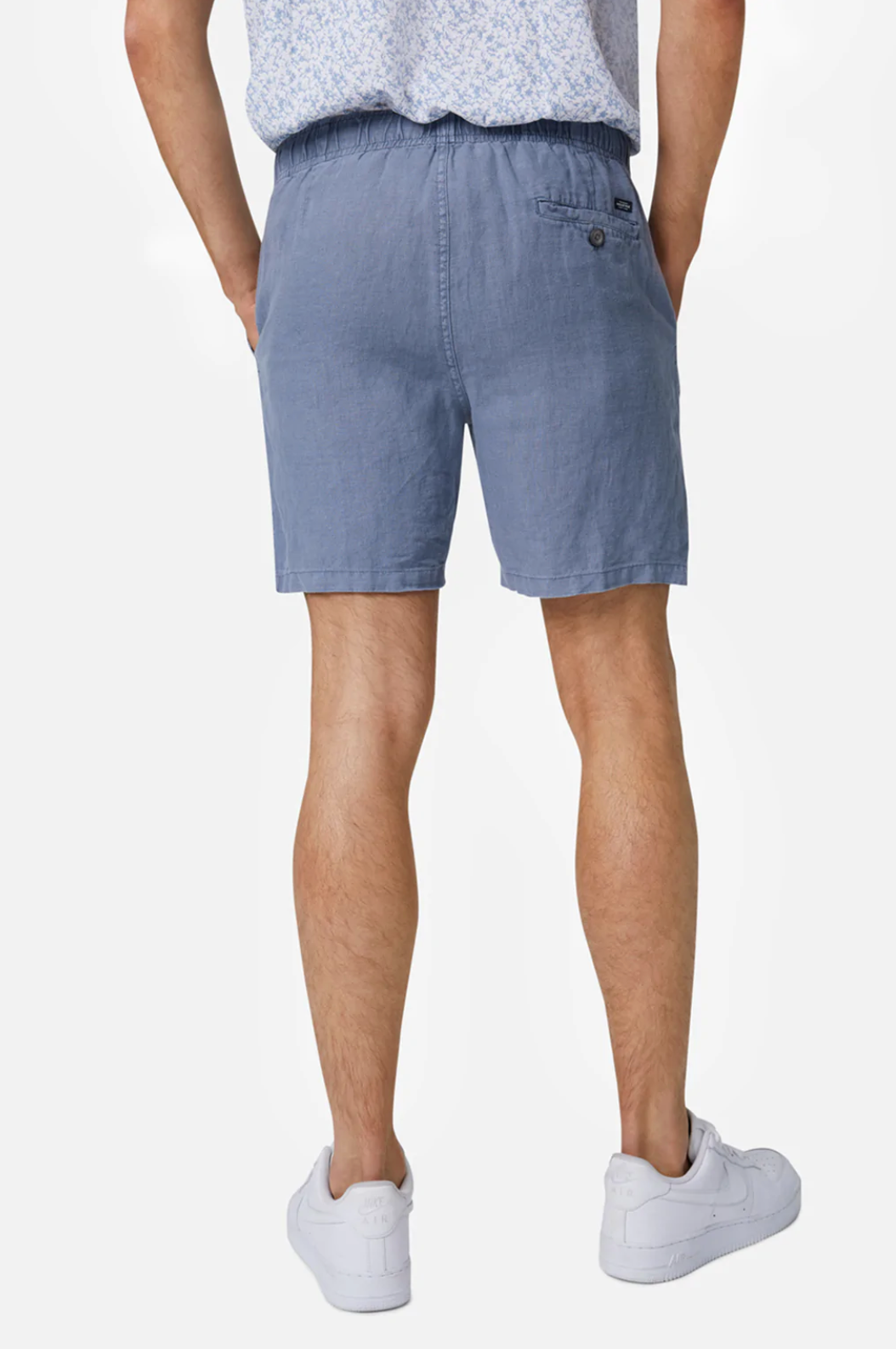A person is seen from the back wearing Industrie The Marina Linen Short in light blue, featuring an elastic drawstring waist and a buttoned back pocket. They're paired with a patterned shirt and white sneakers against a white backdrop.