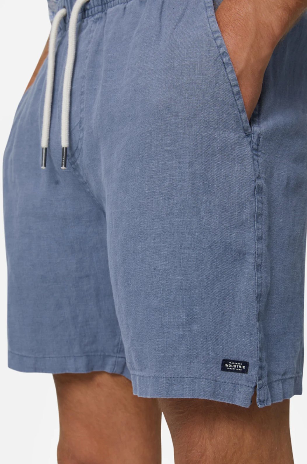 A person wears Industrie The Marina Linen Short in light blue, featuring an elastic drawstring waist and hands in pockets. A small black label is visible near the hem on the left side against a white background.