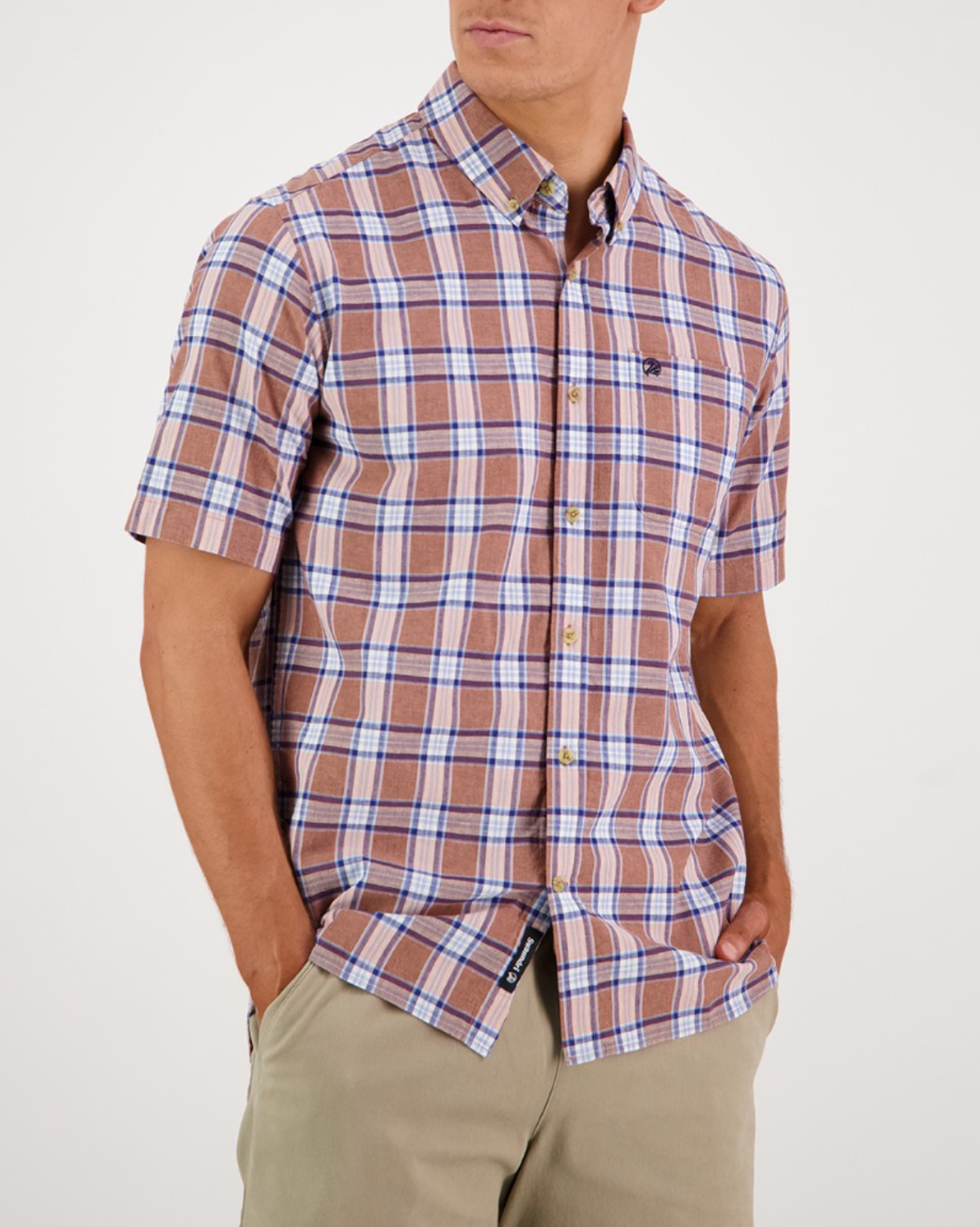 A man stands against a plain white background, hands in the pockets of his beige pants, wearing a Swanndri Kennedy Point Shirt. This tailored-fit shirt features brown plaid with blue and white stripes and is crafted from Cotton and Linen for an elegant look.