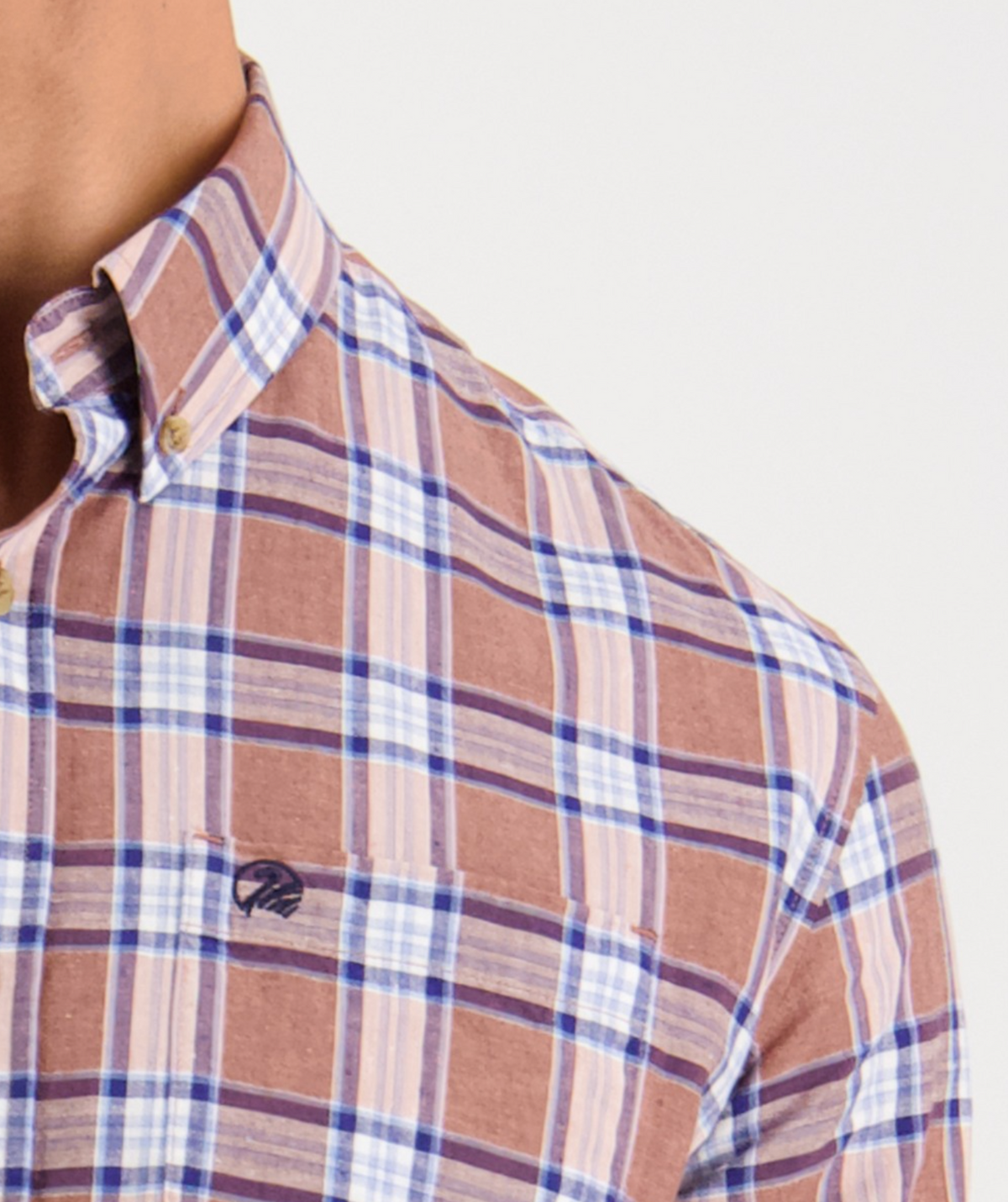 A person in a Swanndri Kennedy Point Shirt with a tailored fit, showcasing a brown, blue, and white plaid pattern. It features an embroidered chest logo and is made from a comfortable cotton-linen blend. The top button is open for casual style against a plain white background.