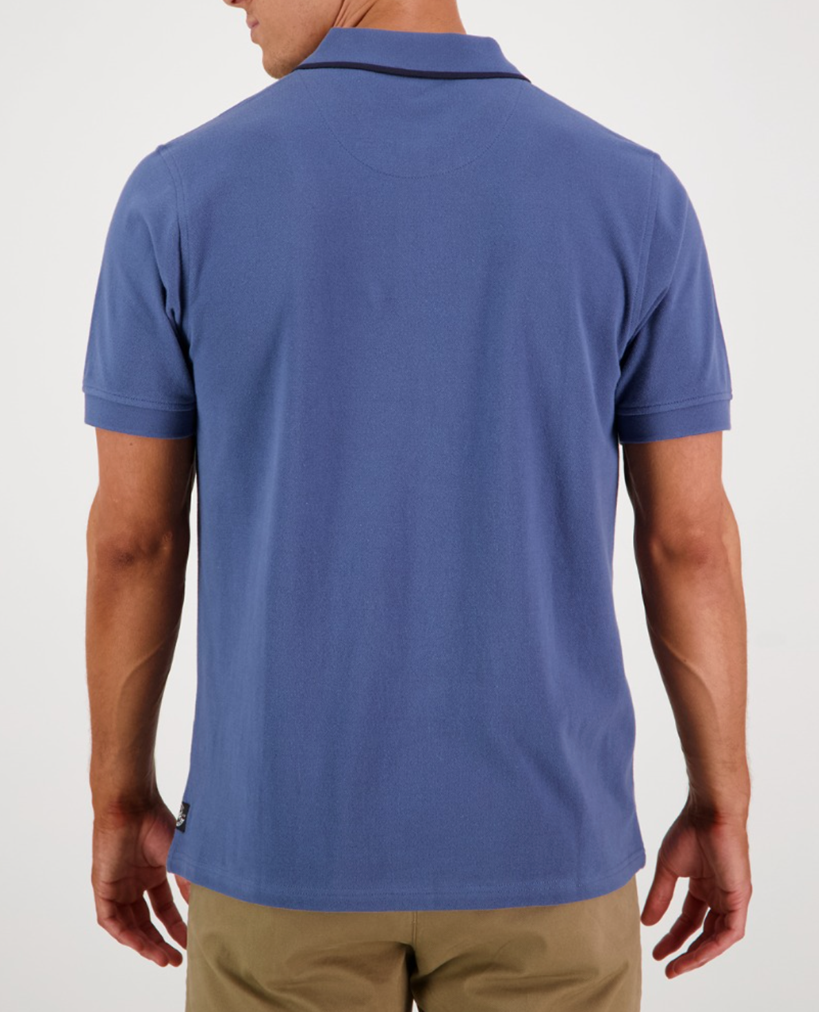 A person in the Swanndri Pinehurst Polo, a blue shirt with an embroidered logo, pairs it with khaki pants. The plain background emphasizes the 100% cotton attire.