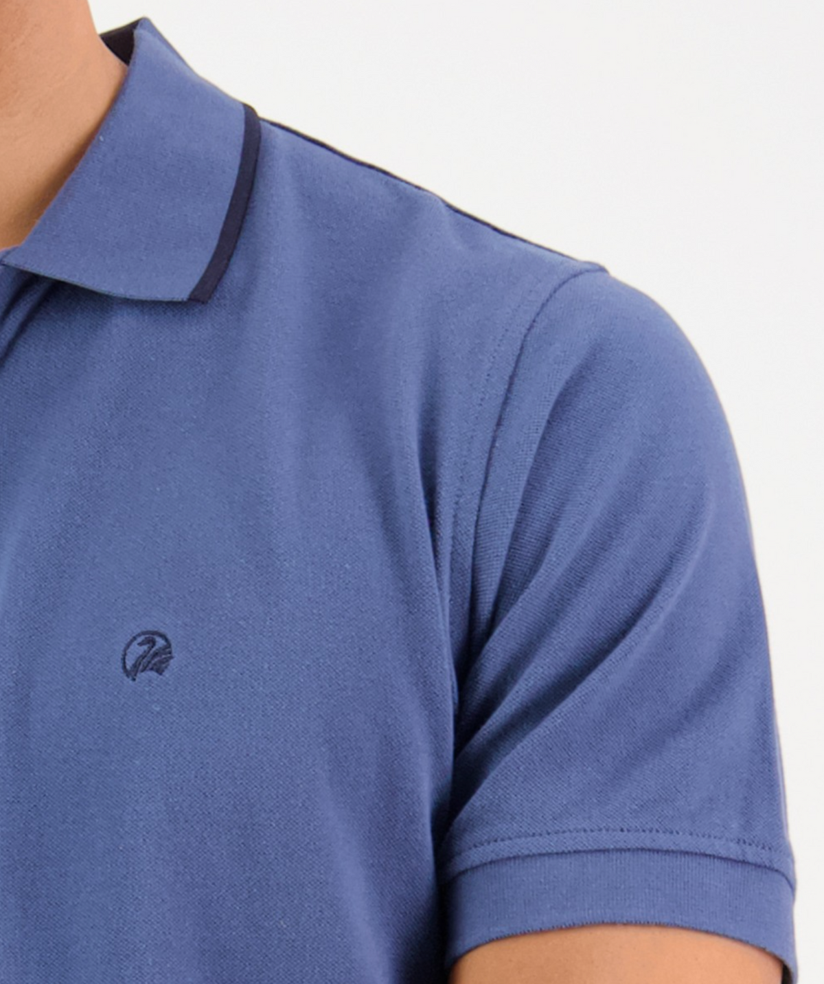 The image shows a close-up of a person wearing the Swanndri Pinehurst Polo, a blue 100% cotton shirt with an embroidered Swanndri logo on the chest and dark trim on the collar. The focus is on the upper torso, including part of the left shoulder.