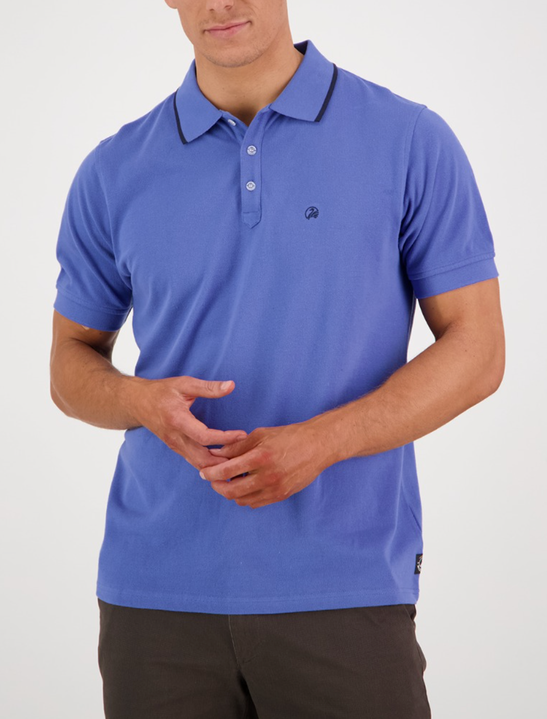 A person models the Swanndri Pinehurst Polo—a 100% cotton, blue shirt with dark trim—against a white background. Their hands are clasped in front, revealing the embroidered logo on the shirt's left side. Dark pants complete the outfit.