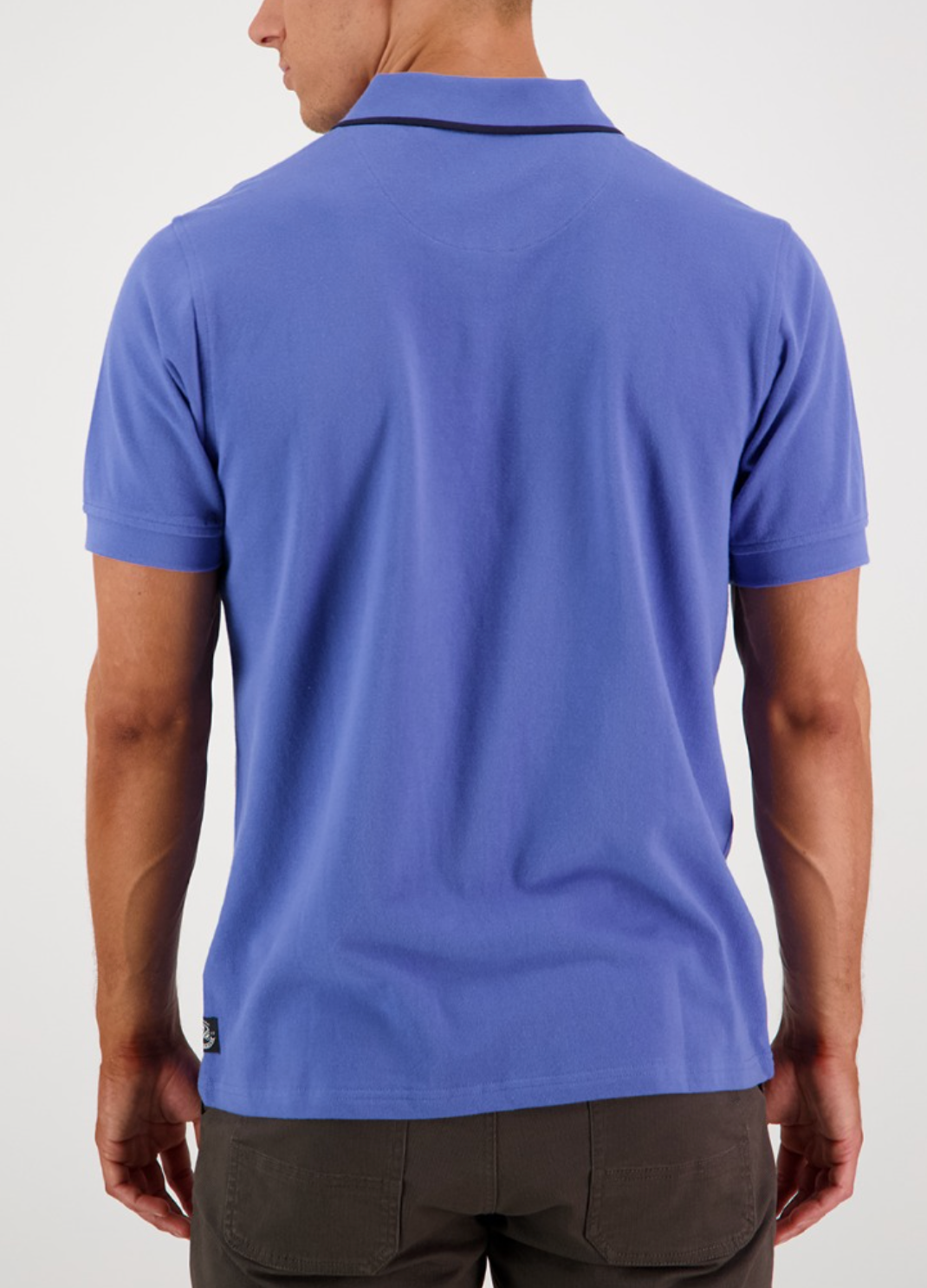 A person wears a Swanndri Pinehurst Polo, featuring the embroidered logo and short sleeves in blue 100% cotton, facing away from the camera. They also wear dark pants against a plain, light background.