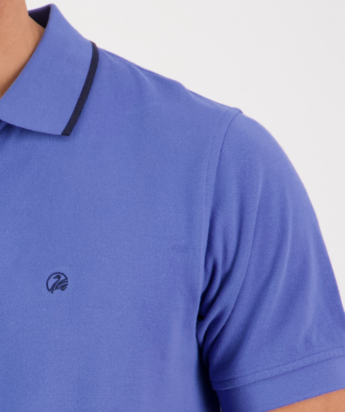 A person wears the Swanndri Pinehurst Polo, a blue shirt with a Swanndri logo on the chest and black collar trim. Made from 100% cotton, it balances comfort and style against a sleek plain white background.