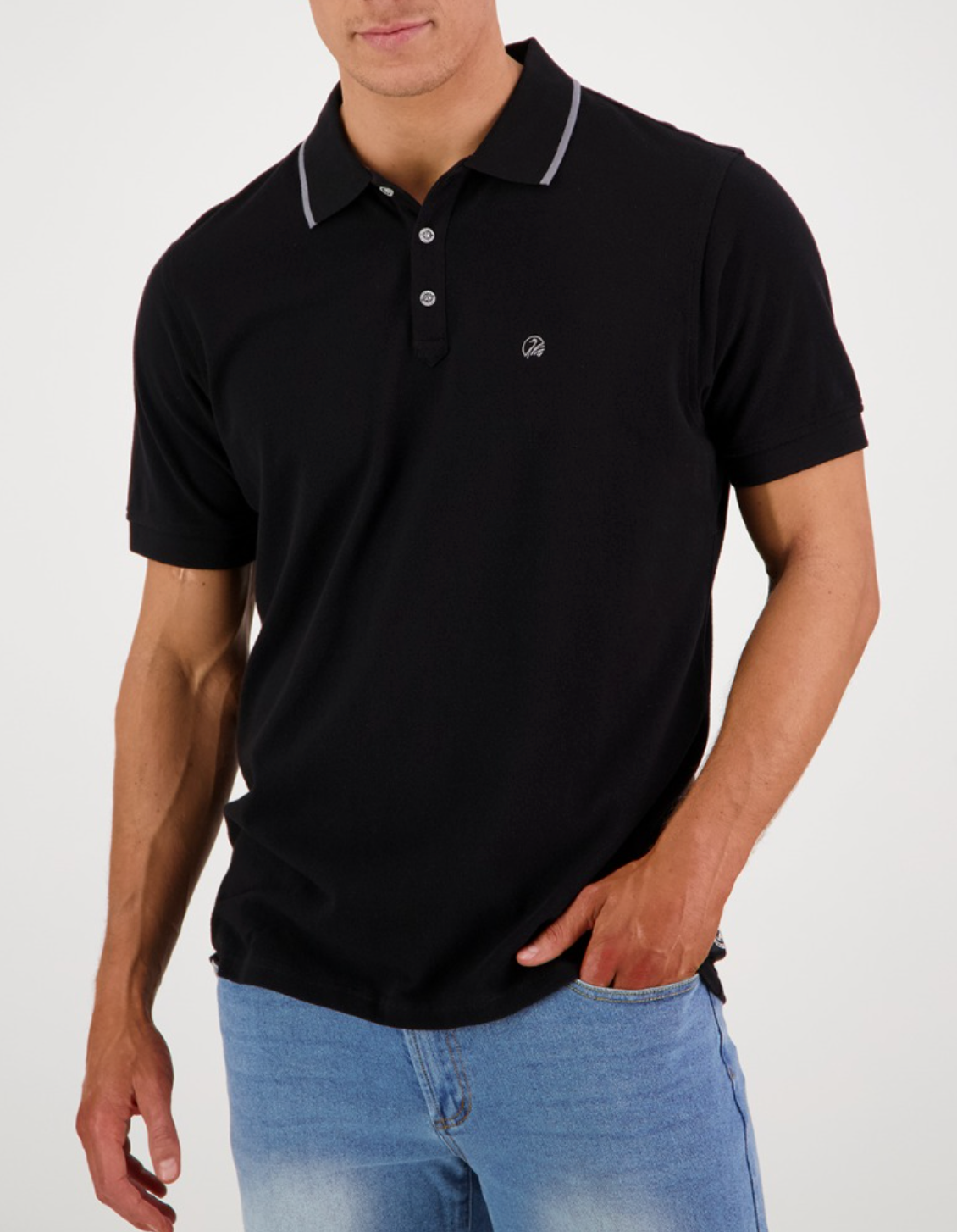 A person in a Swanndri Pinehurst Polo, made of black 100% cotton with dark buttons and a small embroidered logo on the chest, stands with one hand in their blue jeans pocket against a plain white background.