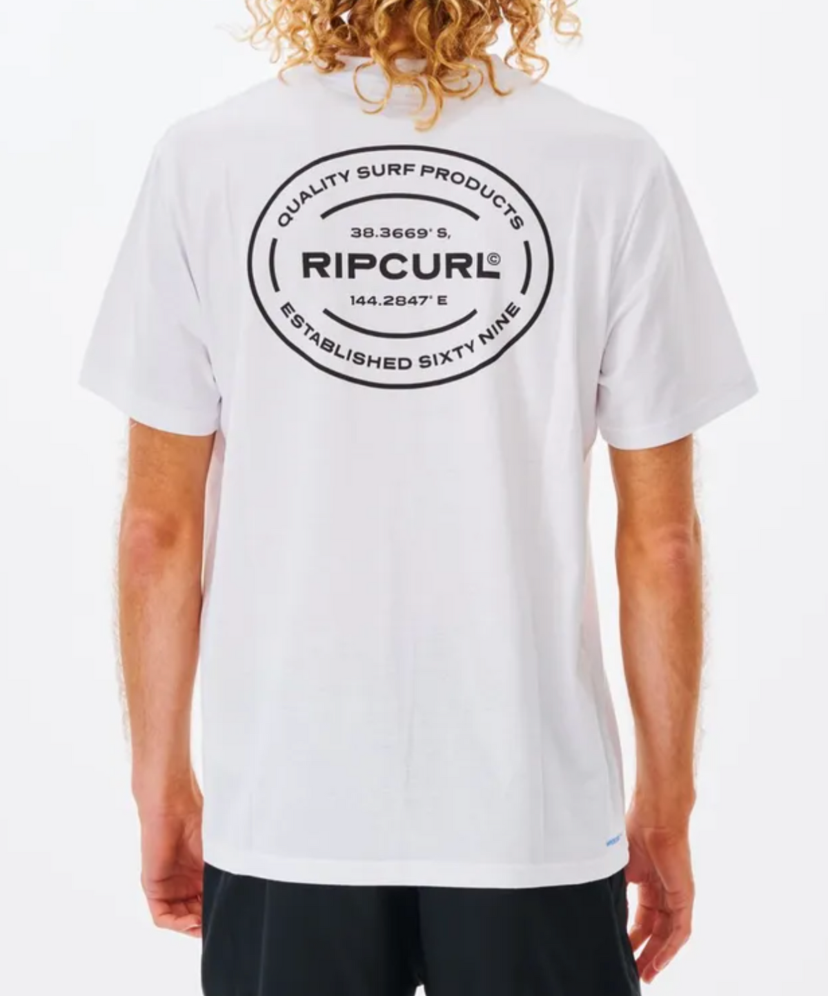 A person with curly blond hair is seen from behind, wearing a white Rip Curl Vaporcool Oval Tee with a large black logo and coordinates on the back, enjoying the moisture-wicking comfort of the fabric.