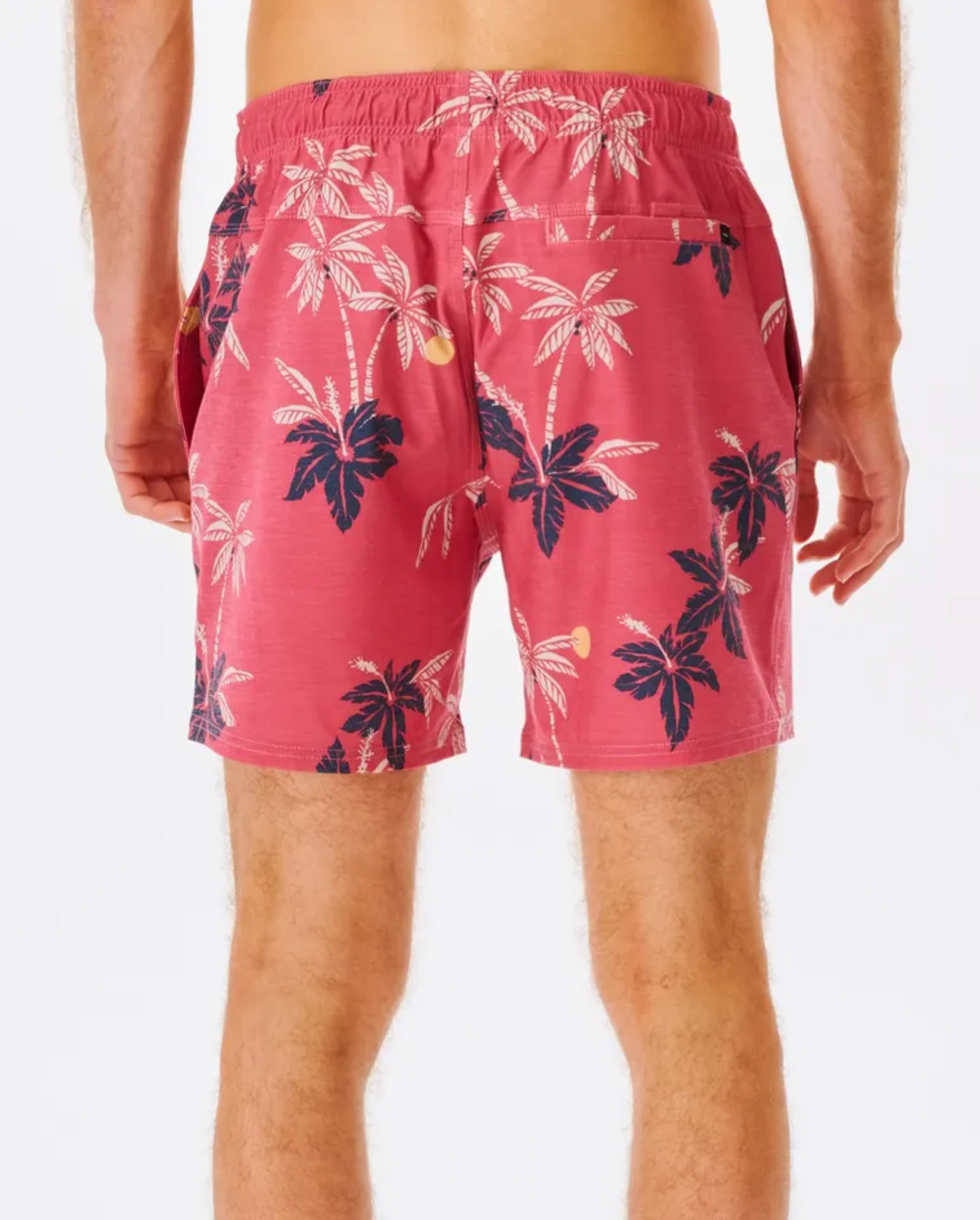The person is wearing Rip Curl Sumatra Volley Shorts, featuring a pink design with dark blue and white palm trees. Made from quick-dry, recycled fabric, these shorts have an elastic waistband and side pockets. They stand against a plain background with legs visible.
