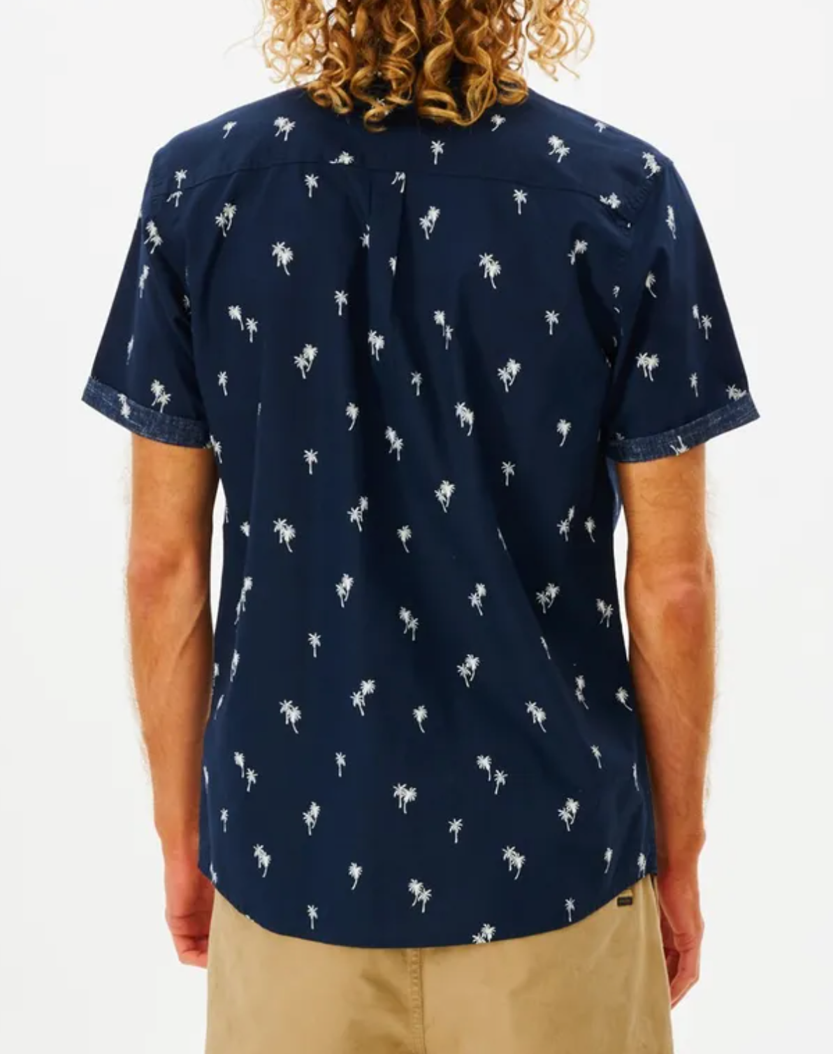 The rear view shows a curly-haired person in a navy blue Rip Curl Paradise Palms Shirt with short sleeves and a palm tree pattern, paired with khaki shorts. The 100% cotton ensemble stands out against a plain white background.