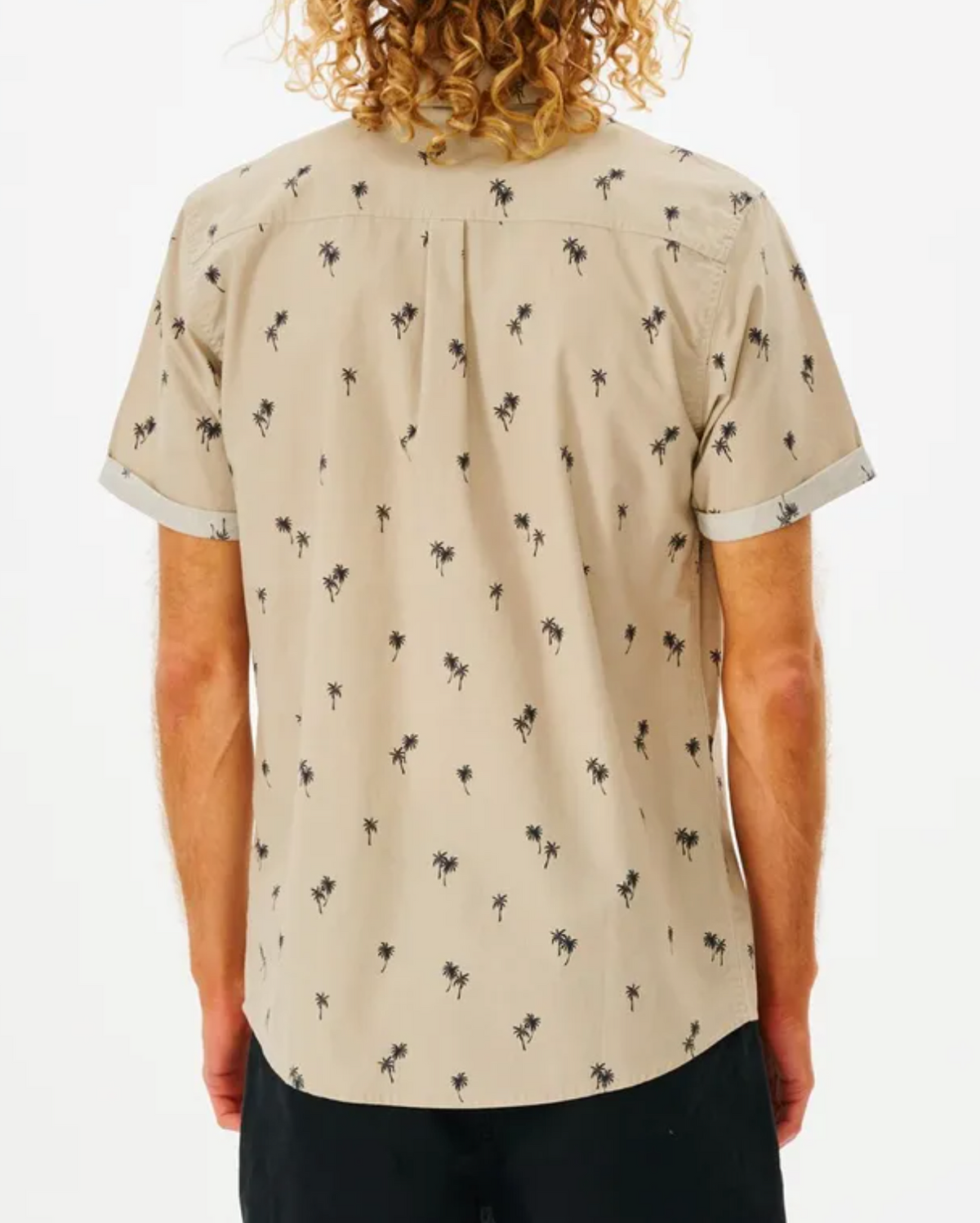 The back view shows a person with curly hair wearing the Rip Curl Paradise Palms Shirt, highlighting black palm tree patterns on beige short sleeves. Made from 100% cotton, it's paired with dark pants against a plain white background.