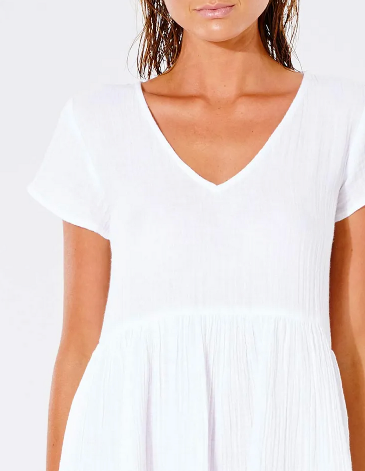 A woman dons a Rip Curl Premium Surf Dress in white, featuring a V-neckline and visible from shoulders to waist. Its short sleeves and textured fabric add comfort. Her wet hair against an off-white background exudes an effortlessly casual vibe.