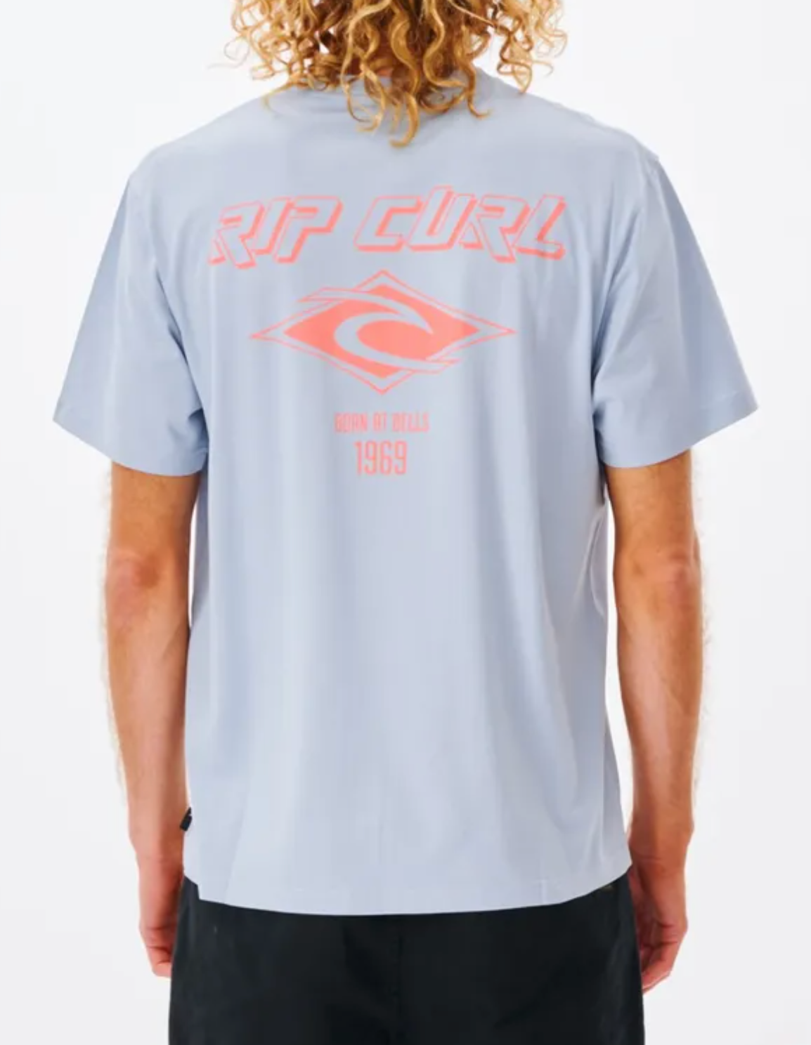 A person with long curly hair wears a relaxed fit light blue Rip Curl Fade Out Icon Tee made from organic cotton, featuring a pink "RIP CURL" logo and "BORN AT BELLS 1969" text design. They're paired with black pants.