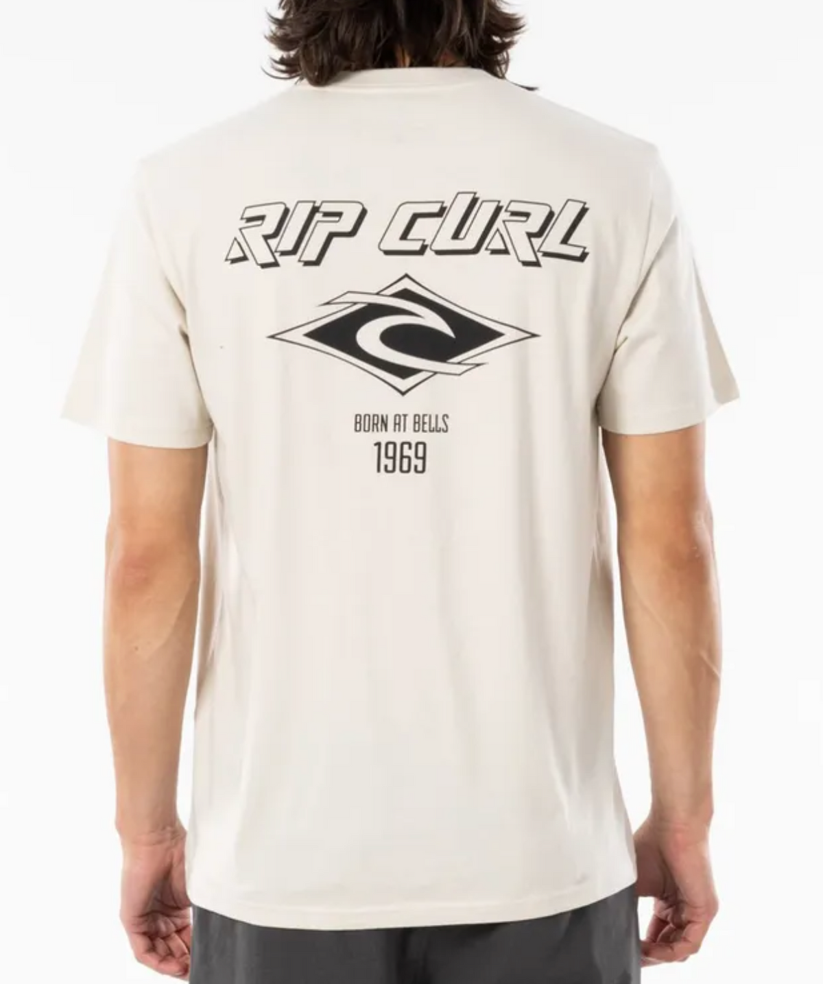 The man is wearing a relaxed fit Rip Curl Fade Out Icon Tee in beige, with "RIP CURL" text, a wave logo, and "BORN AT BELLS 1969" on the back. Made from organic cotton, he's photographed facing away from the camera.
