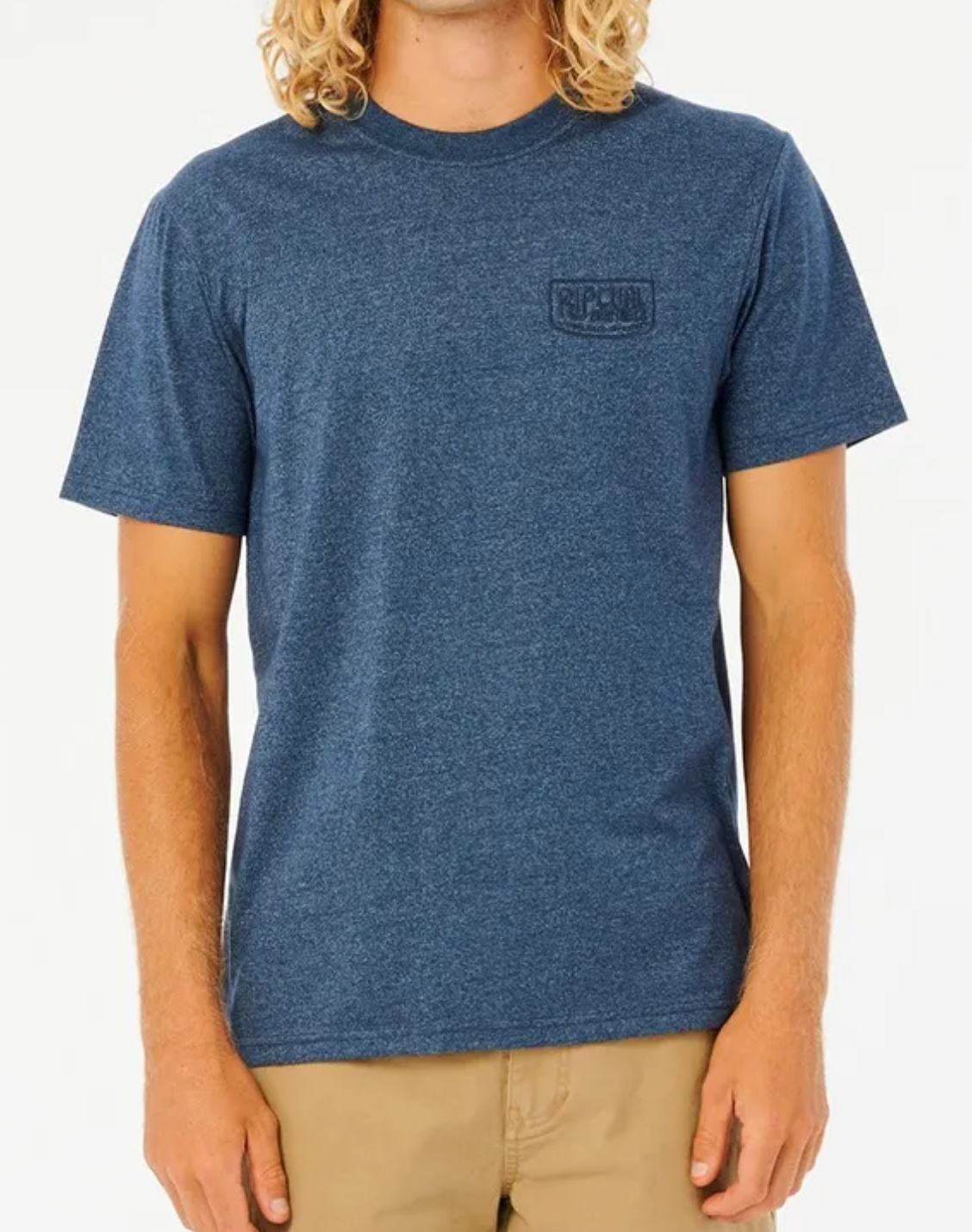 A person models the "Rip Curl Established Tee" featuring its logo, paired with tan pants and their wavy blonde hair against a plain white background. The blue T-shirt, made from a comfy Cotton/Polyester blend, is ideal for a laid-back style.