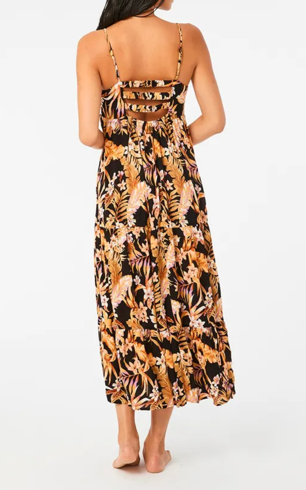 A woman, standing barefoot and facing away, wears the Rip Curl Sunday Swella Midi Dress made of EcoVero fabric. The sleeveless dress features a tropical pattern in orange, pink, and brown with thin straps and cut-out details at the back against a plain white background.
