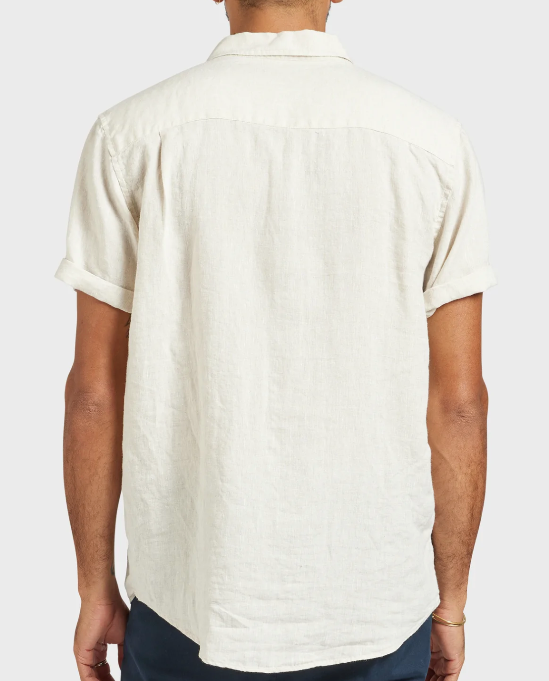 A person in the Academy Brand Hampton Linen Short Sleeve Shirt, light beige, regular fit, seen from the back with rolled cuffs and dark pants, stands against a plain light background.
