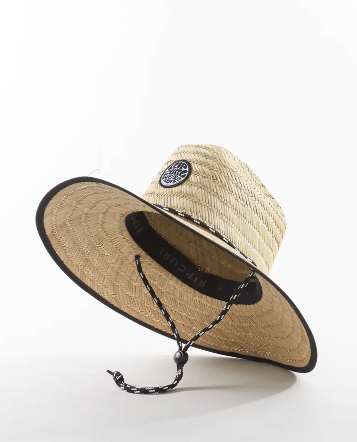 The Rip Curl Icons Straw Hat features a wide-brimmed design with a black band on the edge and a circular front patch. It includes an adjustable chin strap with a black bead, resting on a white surface, angled slightly to the side.