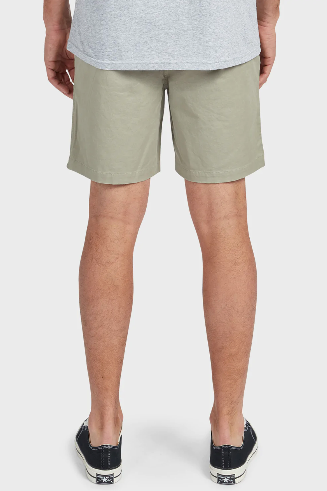 A person is seen from behind wearing Academy Brand Volley Shorts in light gray cotton with an elastic drawstring waist, paired with a gray T-shirt. Their legs are visible, clad in black and white sneakers featuring star designs. The background is plain white.