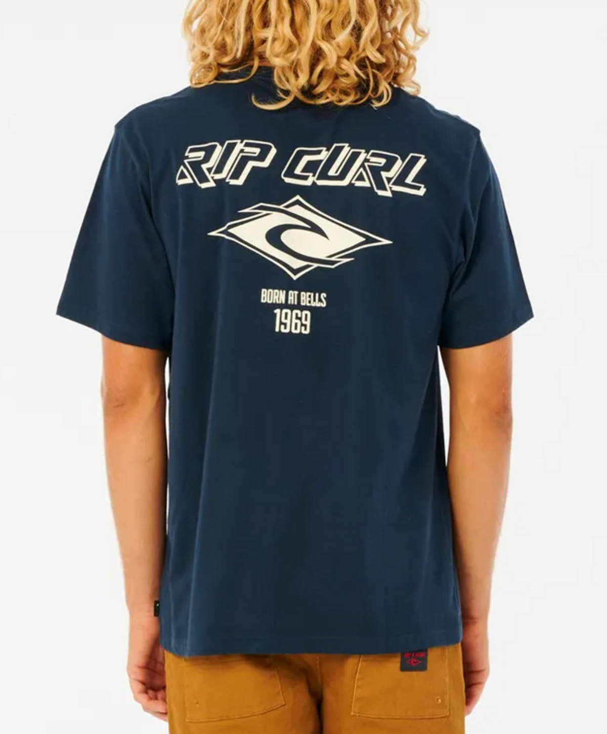 A person with curly blonde hair, facing away, wears a navy blue Rip Curl Fade Out Icon Tee in a relaxed fit made from organic cotton. The tee showcases the iconic "RIP CURL" logo and "Born at Bells 1969" on the back, paired stylishly with brown pants.