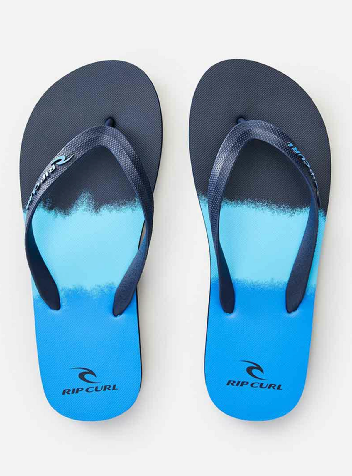 The Rip Curl Big Fade Thongs are blue and black with a gradient design, featuring dark ergonomic molded rubber straps and the logo on the footbed.