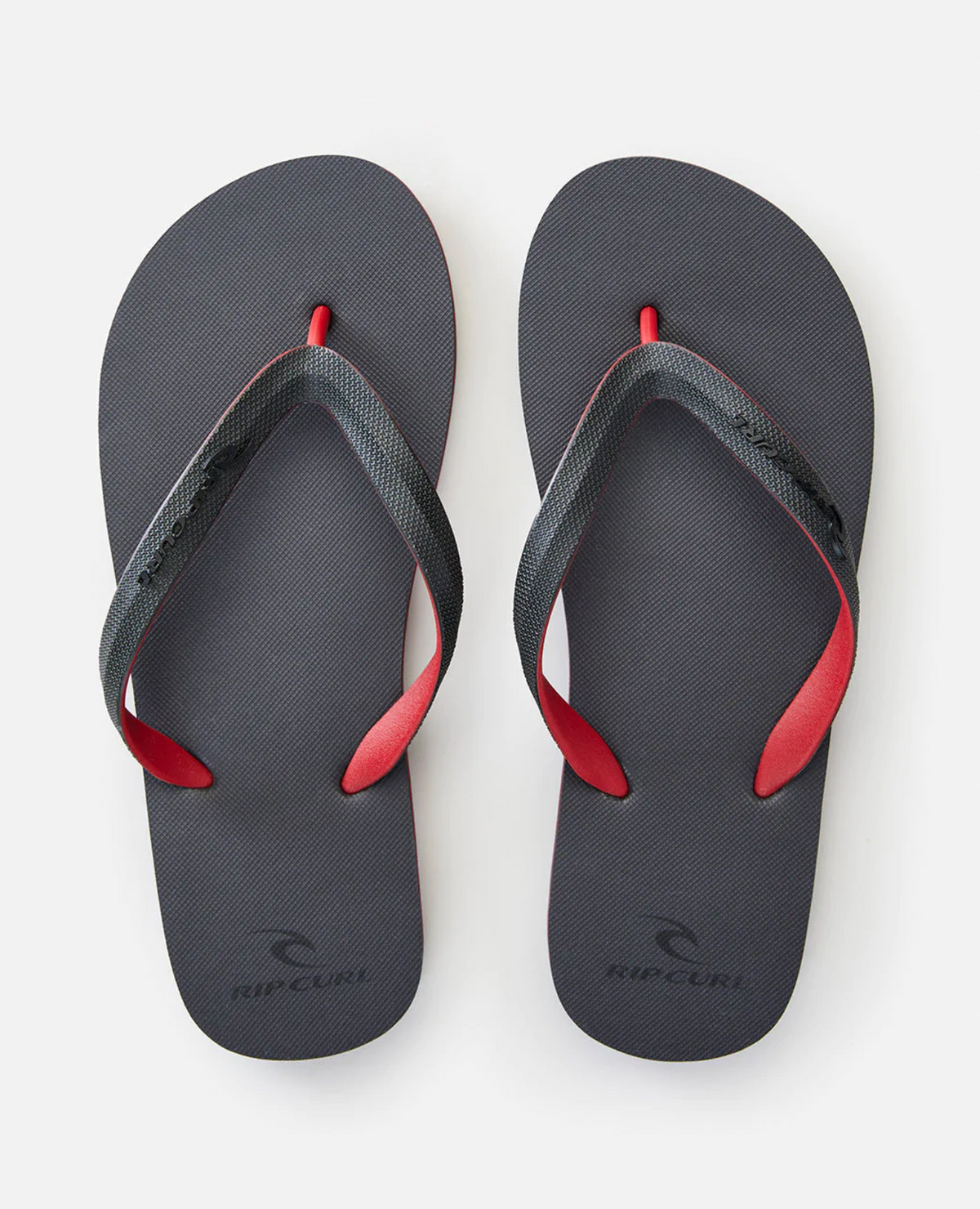 The Rip Curl MC 2 Tone Thongs, featuring black straps with red accents and a thermo rubber compound for comfort, prominently display the brand name on straps and soles. Presented on a sleek white background, they capture attention with their stylish details.