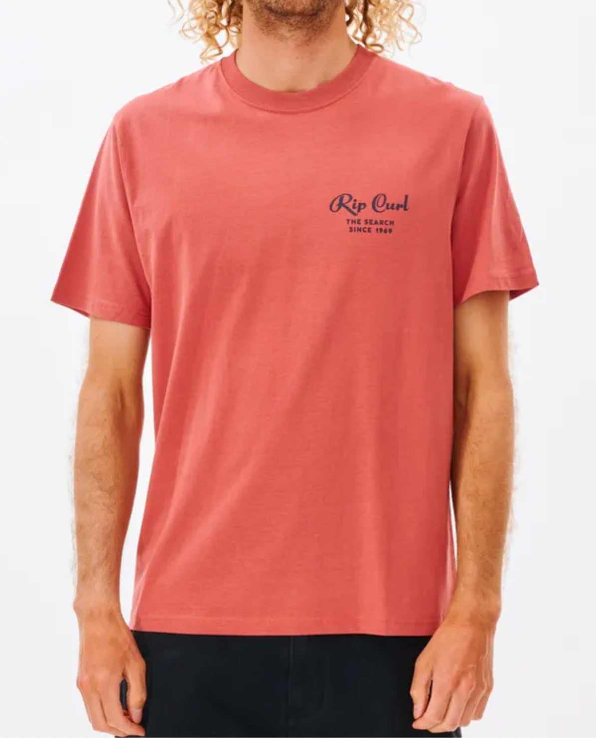 A curly-haired person wears a comfy red Rip Curl Twist Tee featuring "The Search Since 1969," its simple design popping against the plain white backdrop.