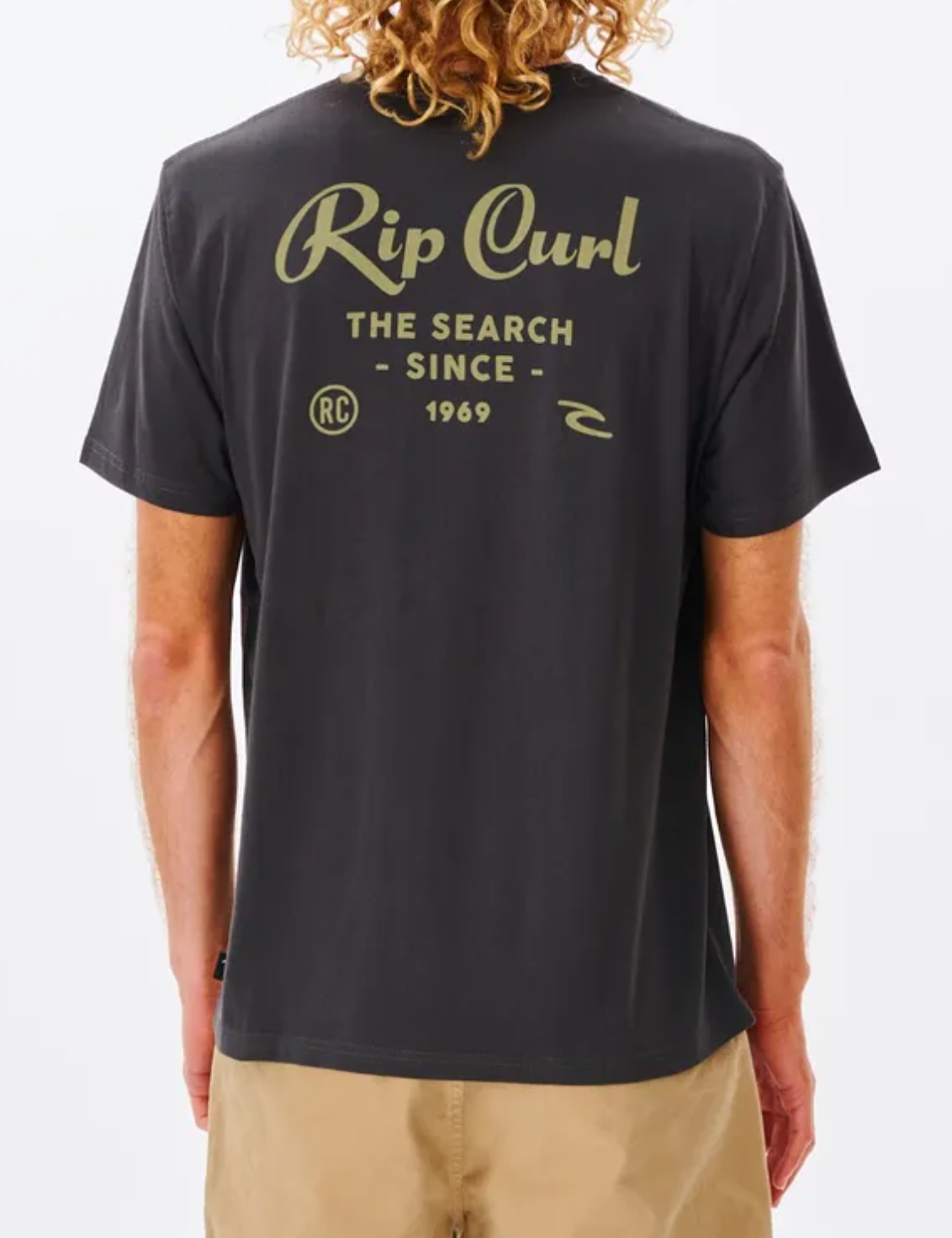A person with curly hair wears a Rip Curl Twist Tee in black, featuring "The Search Since 1969" and olive green circular logos on the back. Paired with khaki shorts, this look embodies effortless style.