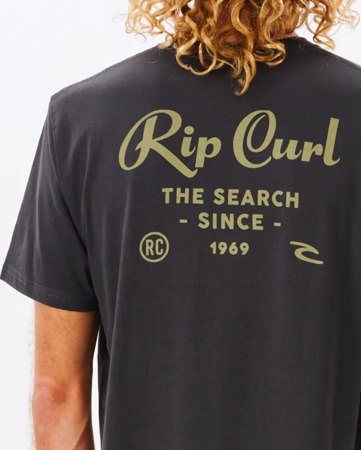 A person with curly hair wears the Rip Curl Twist Tee in black, featuring beige script on the back that reads "The Search Since 1969." A small logo and "RC" initials offer extra detail to this casual, comfortable tee.