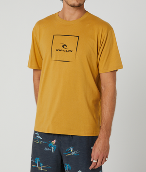 A man wears a mustard yellow Rip Curl Corp Icon Tee (100% cotton, black logo) and navy blue shorts with a tropical beach pattern. The background is plain white.