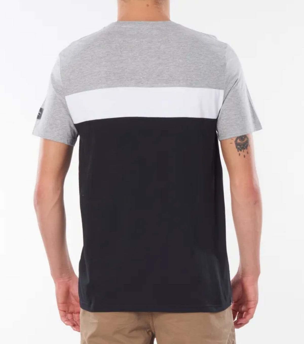 A person wearing a Rip Curl Undertow Panel Tee with gray, white, and black color blocking and beige shorts stands against a plain white background. Their tattooed left arm adds flair as they face away from the camera.