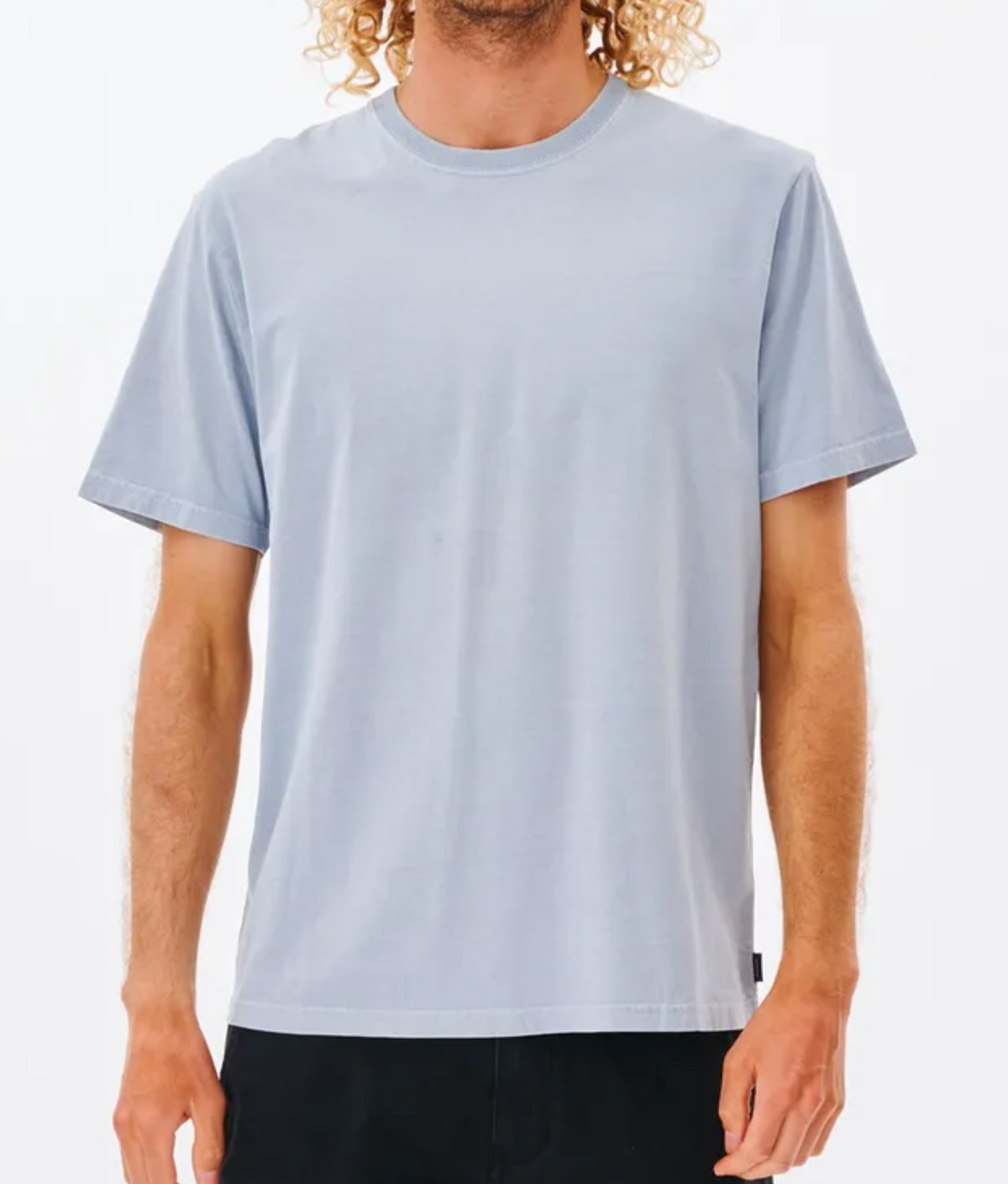 A person with curly blonde hair wears a light blue, short-sleeved Rip Curl Plain Wash Tee, made from 100% cotton. The face is not visible. The individual is standing against a plain white background and is also wearing dark pants. This comfortable tee showcases a relaxed fit, perfect for everyday wear.