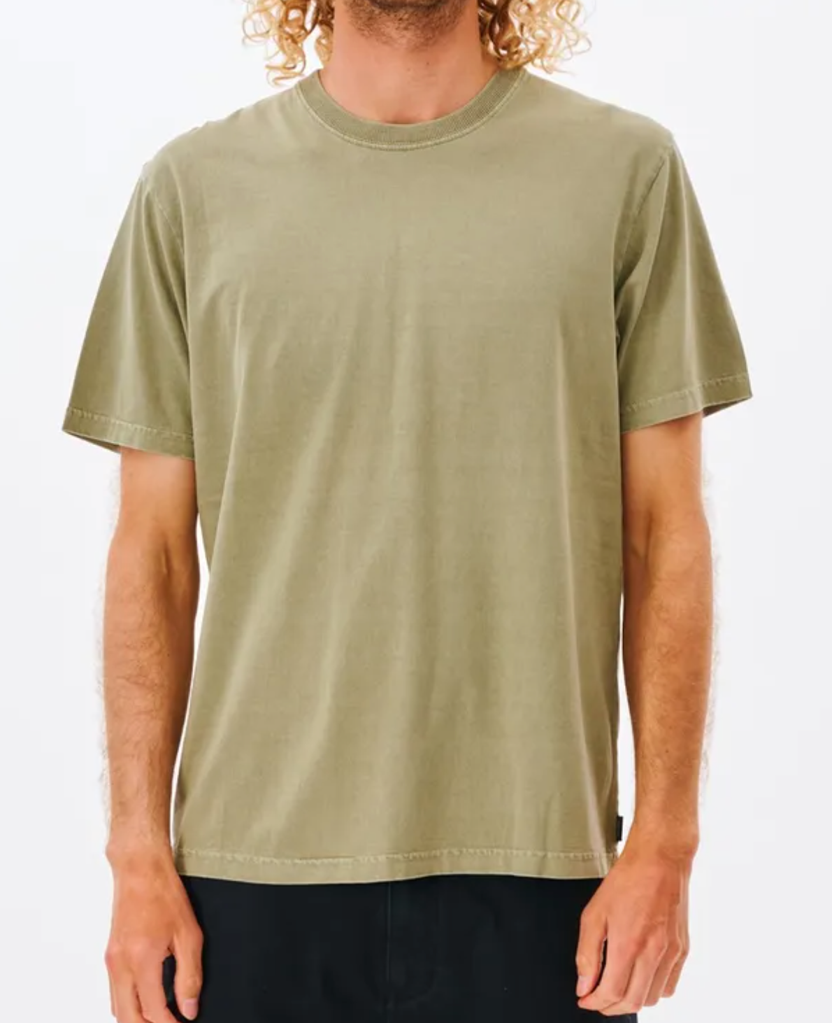 Man in a Rip Curl Plain Wash Tee and black pants standing against a white background. The person has long curly hair cascading over the shoulders and is standing with arms relaxed at sides. The comfortable olive-green tee features short sleeves and a crew neck for a relaxed fit.