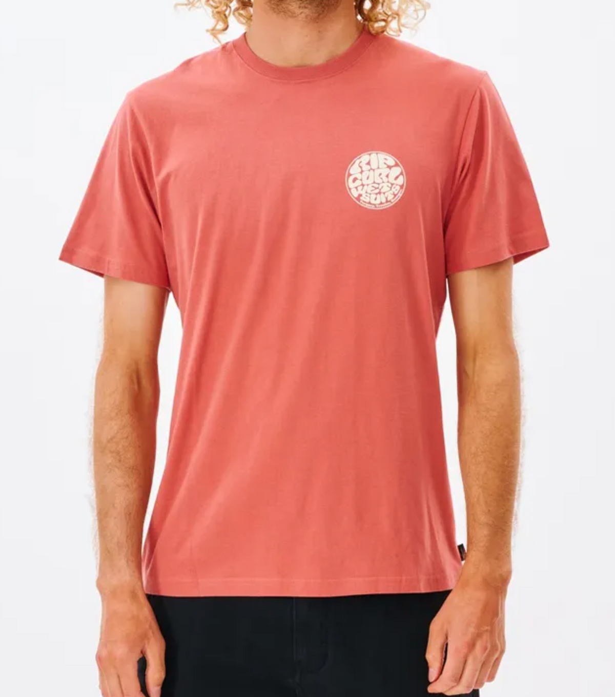 A person with curly blonde hair is wearing a comfortable Rip Curl Wetsuit Icon Tee, a red short-sleeved, 100% cotton shirt with a white circular logo that says "Surf Coast Co." The logo is printed on the left chest, and the person is standing against a plain white background.