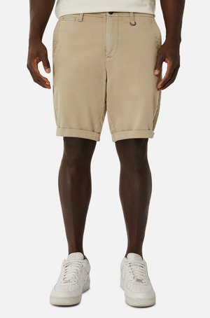 Wearing Industrie The Rinse Drifter Short in beige, a person stands with legs slightly apart against a plain white backdrop, paired with a relaxed-fit white top and white sneakers. Only the lower half of the body is visible.