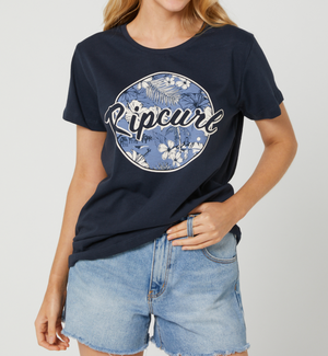 Someone wearing a Rip Curl Beachside Standard Tee in navy blue, made from 100% cotton with a floral graphic, paired with denim shorts, stands against a plain light background.