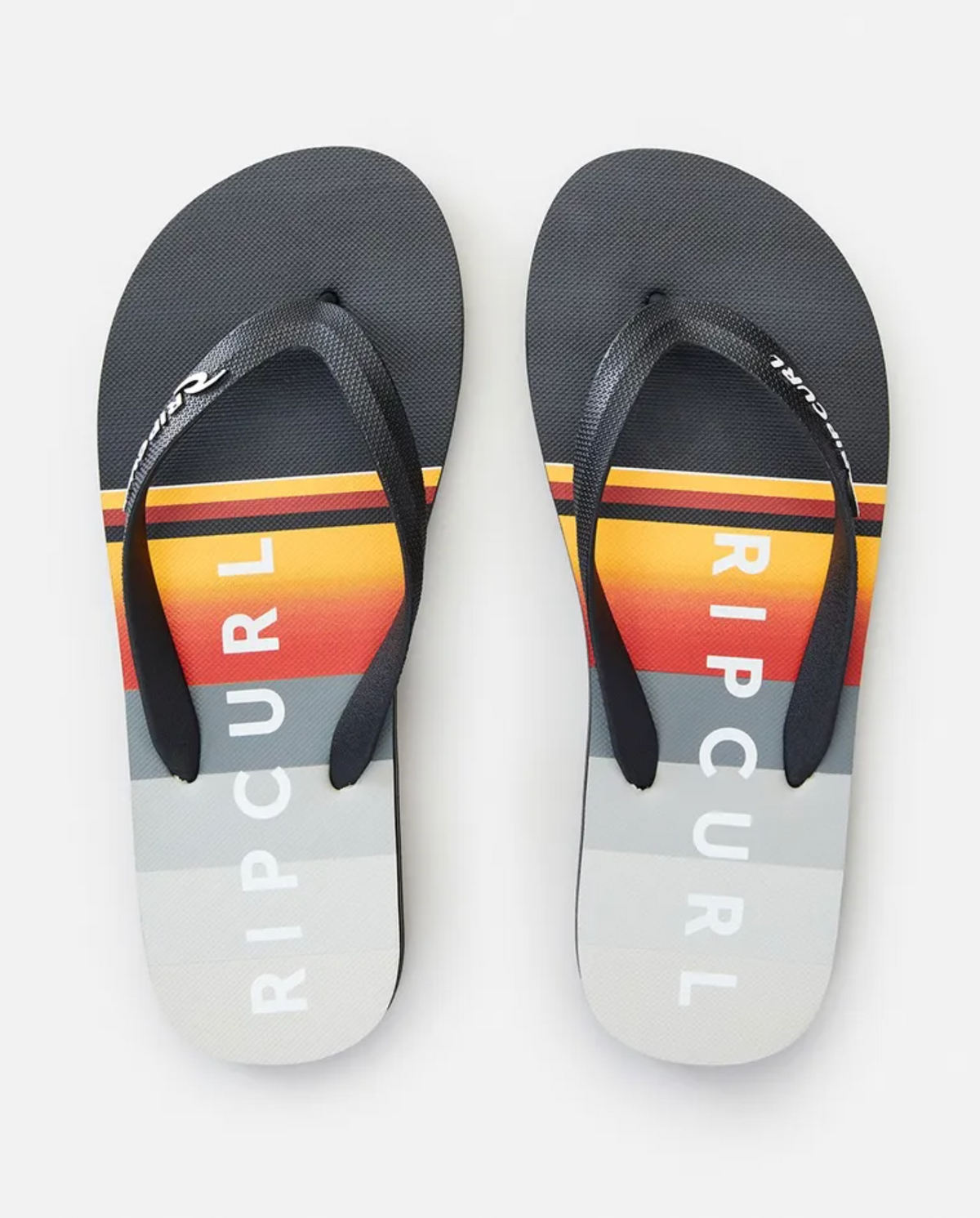 The Rip Curl Daybreak Thongs feature ergonomic molded rubber straps and premium EVA rubber footbeds. The footbeds display a gradient striped pattern in black, yellow, red, and various shades of grey, with "RIP CURL" prominently printed in white.