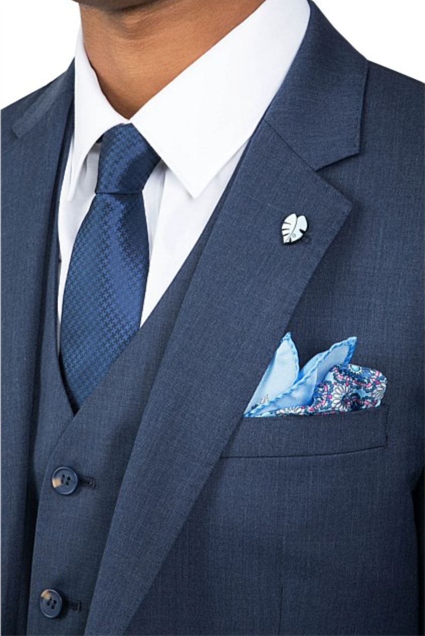 Close-up of someone wearing the Uberstone Troy Jacket, a dark blue slim fit suit with a white shirt and blue tie, featuring a leaf-shaped lapel pin and patterned blue pocket square.