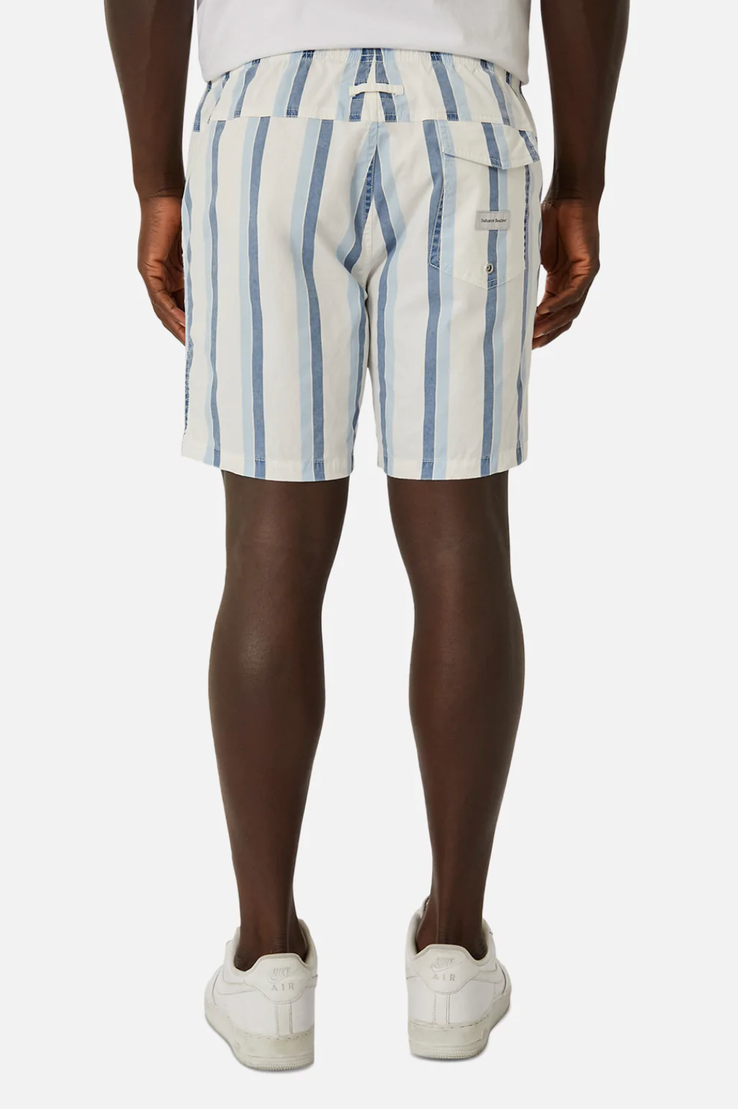 A person is wearing Industrie The Rubio Bahama Short in white with vertical blue stripes and an elastic waist, paired with a white shirt and sneakers. The photo highlights the branding on the back pocket against a plain white background.