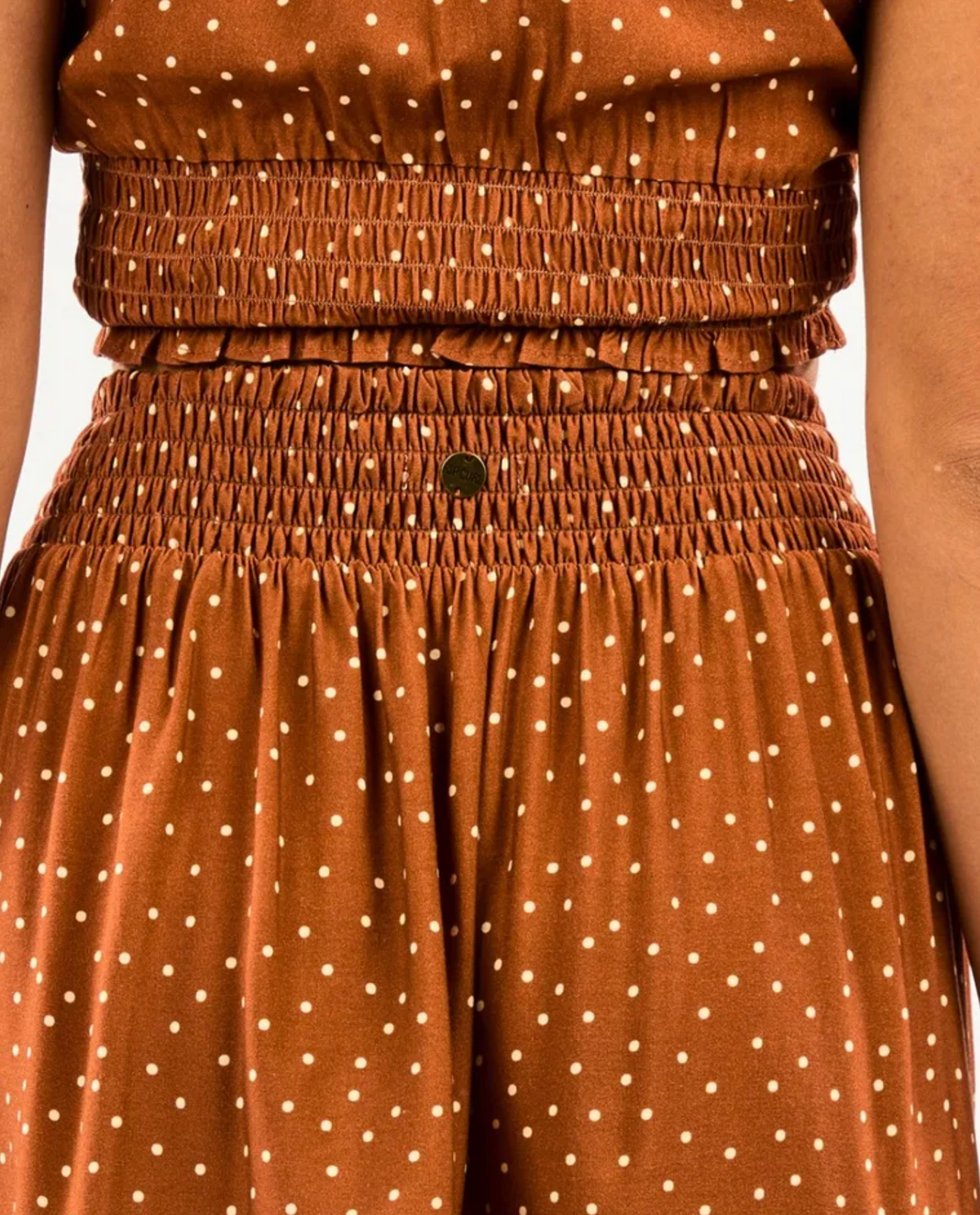 Close-up of a person in the Rip Curl Dreamer Spot Mini Skirt, featuring a smocked waist and chest design. The textured fabric with evenly spaced brown polka dots adds a playful touch. Slightly visible arms complete this chic ensemble.