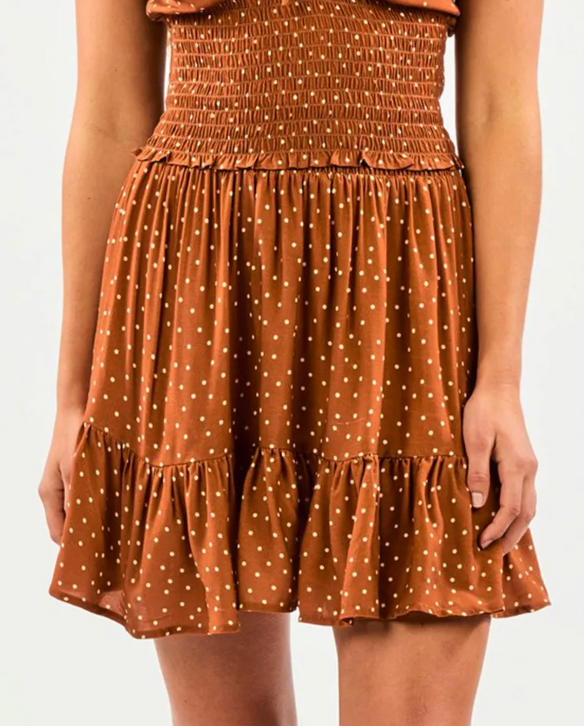 A person wearing a Rip Curl Dreamer Spot Mini Skirt in brown with white polka dots, featuring a smocked waist and ruffled hem. The outfit has a breezy, summer flair that complements the neutral background and exudes a casual, Rip Curl-inspired vibe.