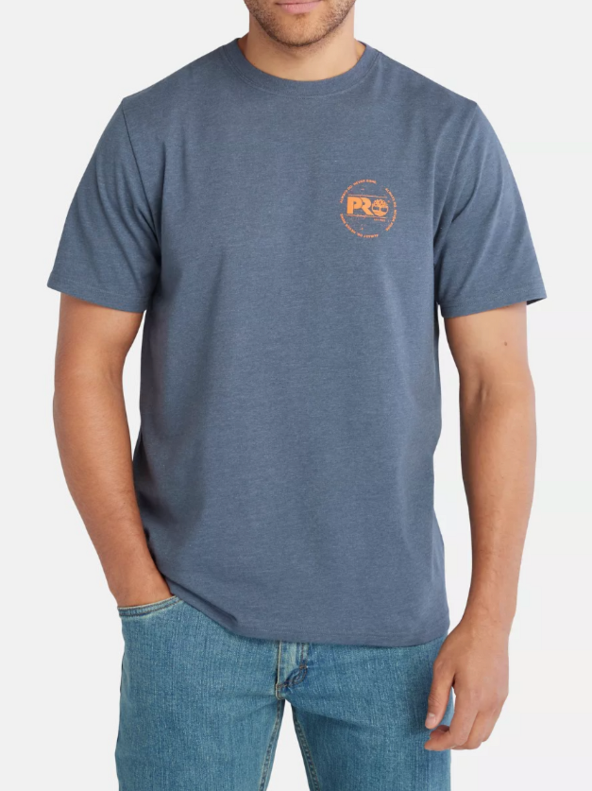 A person in a blue Timberland Pro Base Plate A.D.N.D Graphic Tee, made from recycled polyester with a small orange logo on the left chest, paired with blue jeans. Their hands are in their pockets against a plain white background, exuding casual flair.