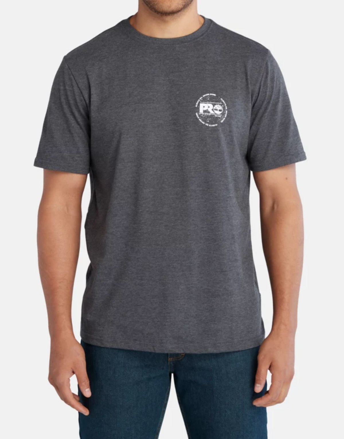 Dressed in blue jeans, a person sports a Timberland Pro Base Plate A.D.N.D Graphic Tee made from recycled polyester in dark gray. The tee, featuring a white chest logo, short sleeves, and crew neck, is moisture-wicking and contrasts the plain white background.