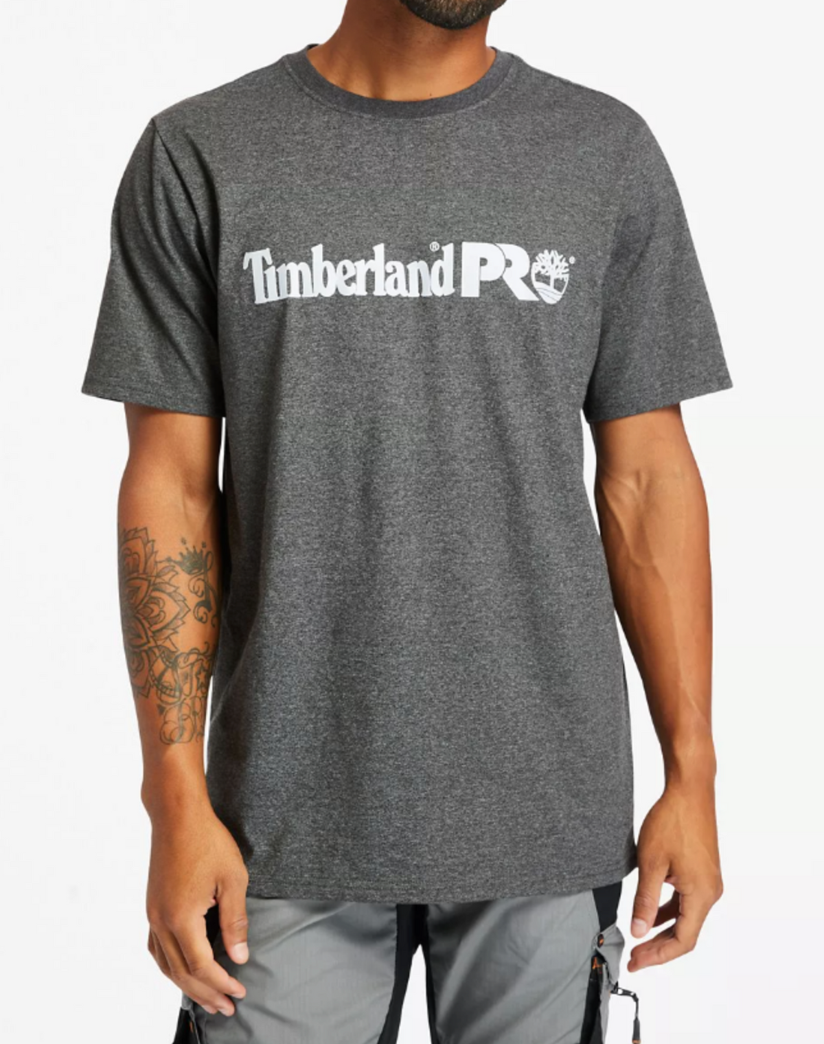 A person is wearing a gray Timberland Pro Base Plate Logo Tee with white lettering and an original fit. The wicking fabric highlights their tattooed left forearm, paired seamlessly with light gray pants against a simple white backdrop.