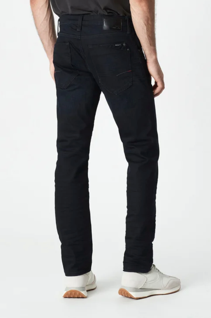 A person wearing Mavi Marcus Slim Straight Jeans in black and white sneakers with tan soles stands with their back to the camera. The jeans have visible stitching and a back pocket, set against a plain white background.